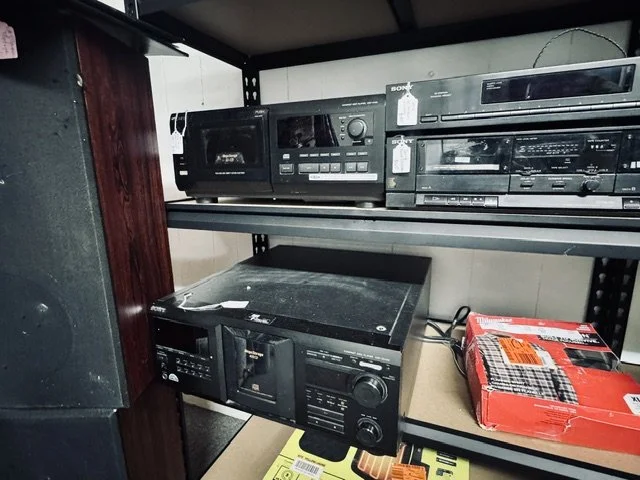 A black electronic stereo receiver on a metal shelf with other audio equipment and a red boxed item nearby, with a wooden vertical support on the left.
