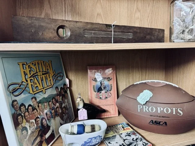 A wooden shelf displays a compilation of nostalgic items including a vintage 'Festival of Faith' poster, a small framed picture of a woman, a ceramic figure, a large fake football labeled 'Prop Bets,' and a few magazines.