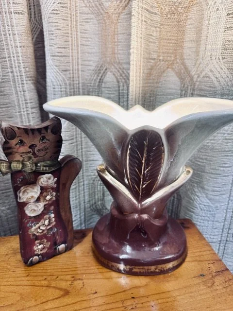 Decorative ceramic vase with wide opening and leaf design, placed on a wooden surface, with a cat-themed decorative figure in the background.