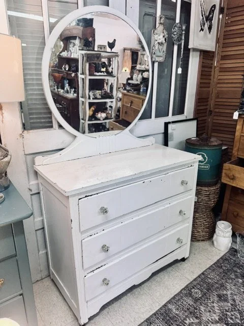 White dresser with a mirror on top in an antique store, surrounded by other vintage furniture and decor.