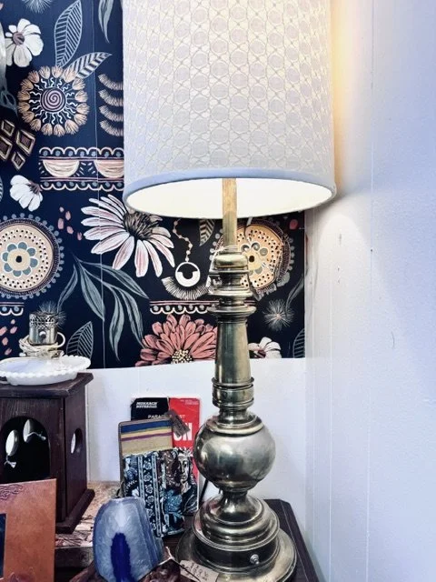 A table lamp with a metallic base and a large white shade, located on a dark wooden surface against a colorful floral and geometric patterned wallpaper background.