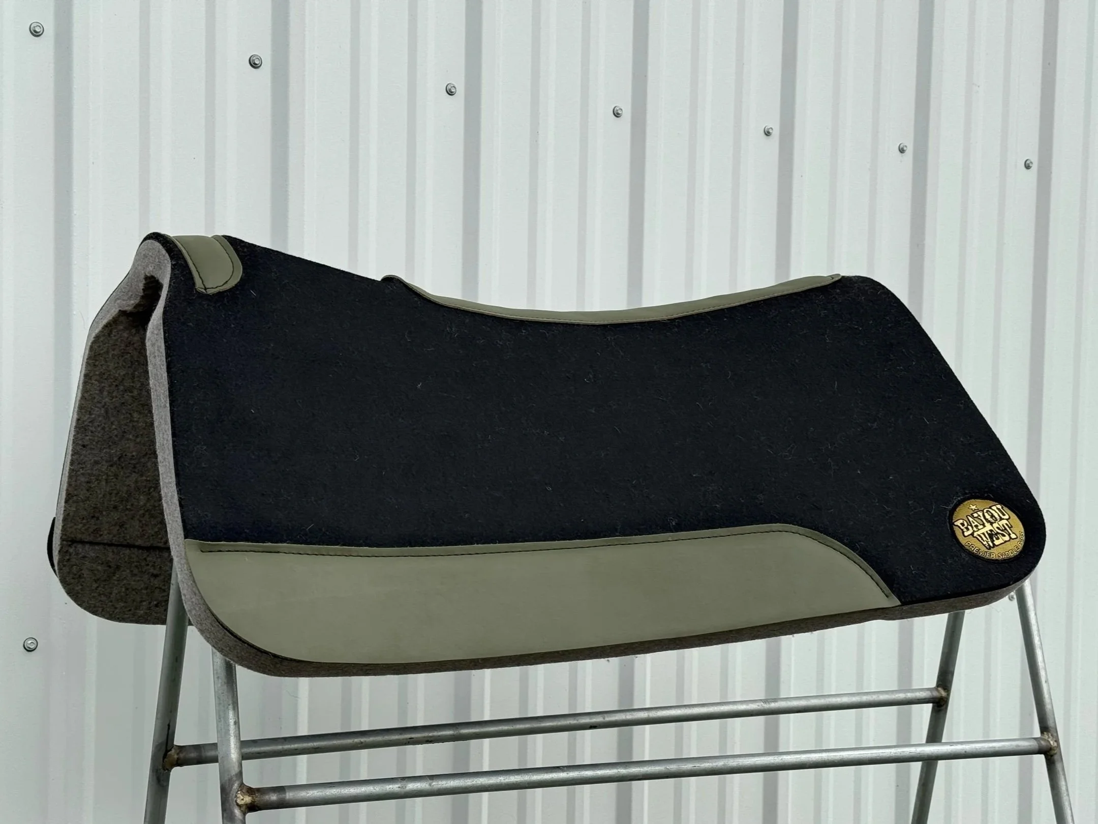 A Black and gray horse saddle with a round golden badge, placed on a metal stand against a white corrugated metal wall.