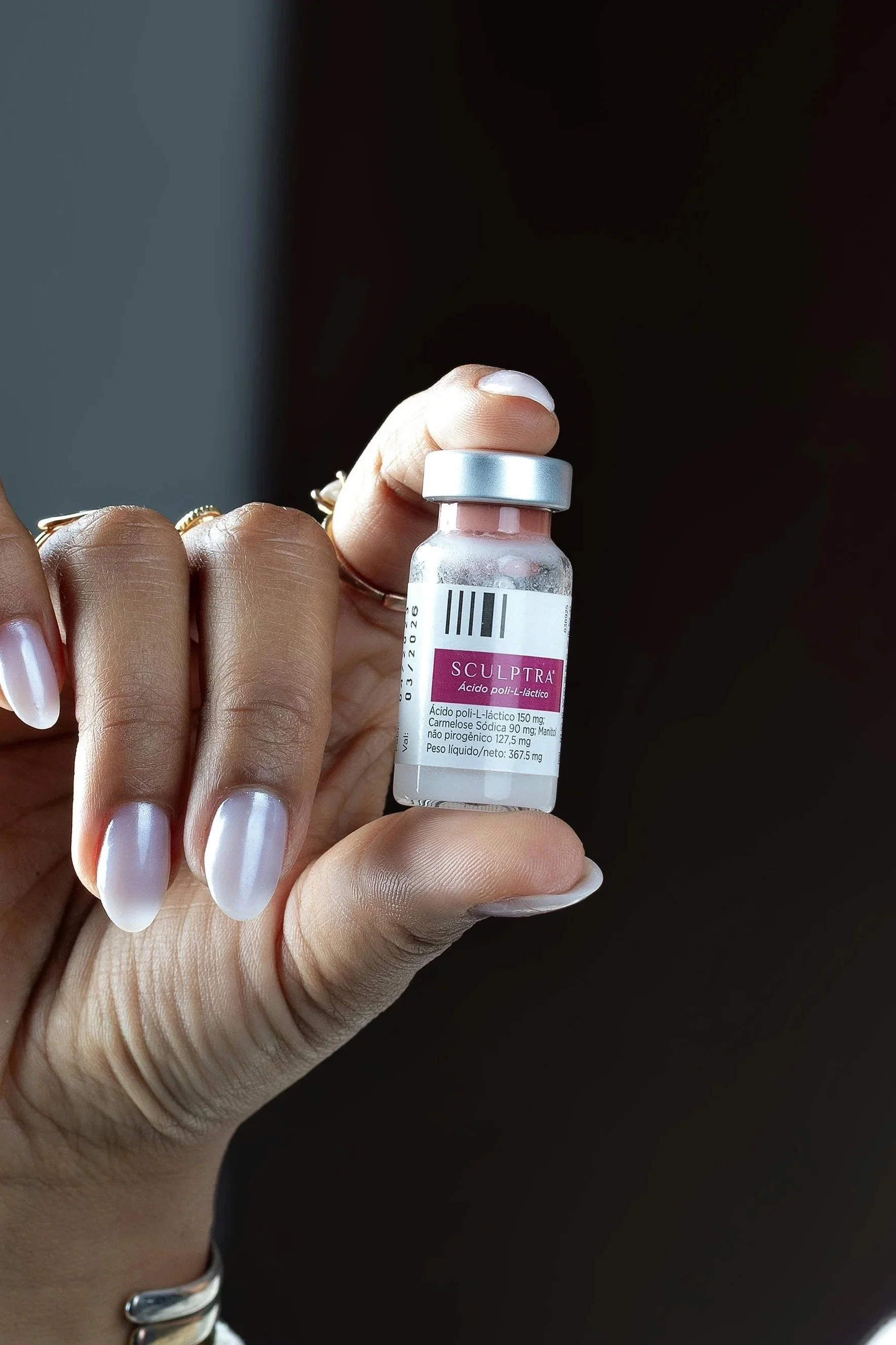 A person holding a small glass vial labeled 'Sculptra' with medication information in Spanish, including active ingredients and dosages.