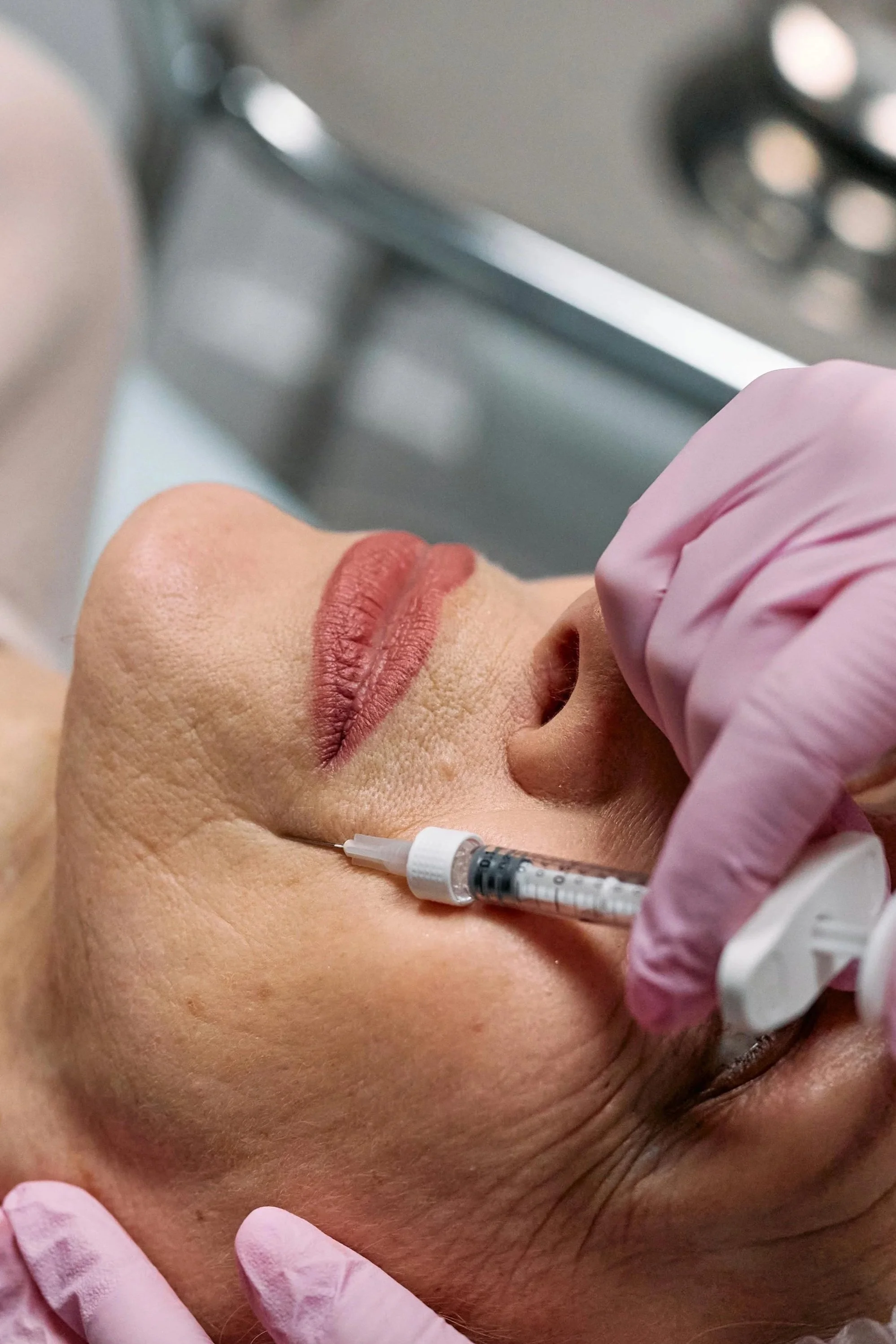 A woman receiving an injection in her cheek from a medical professional wearing pink gloves, in a clinical setting.