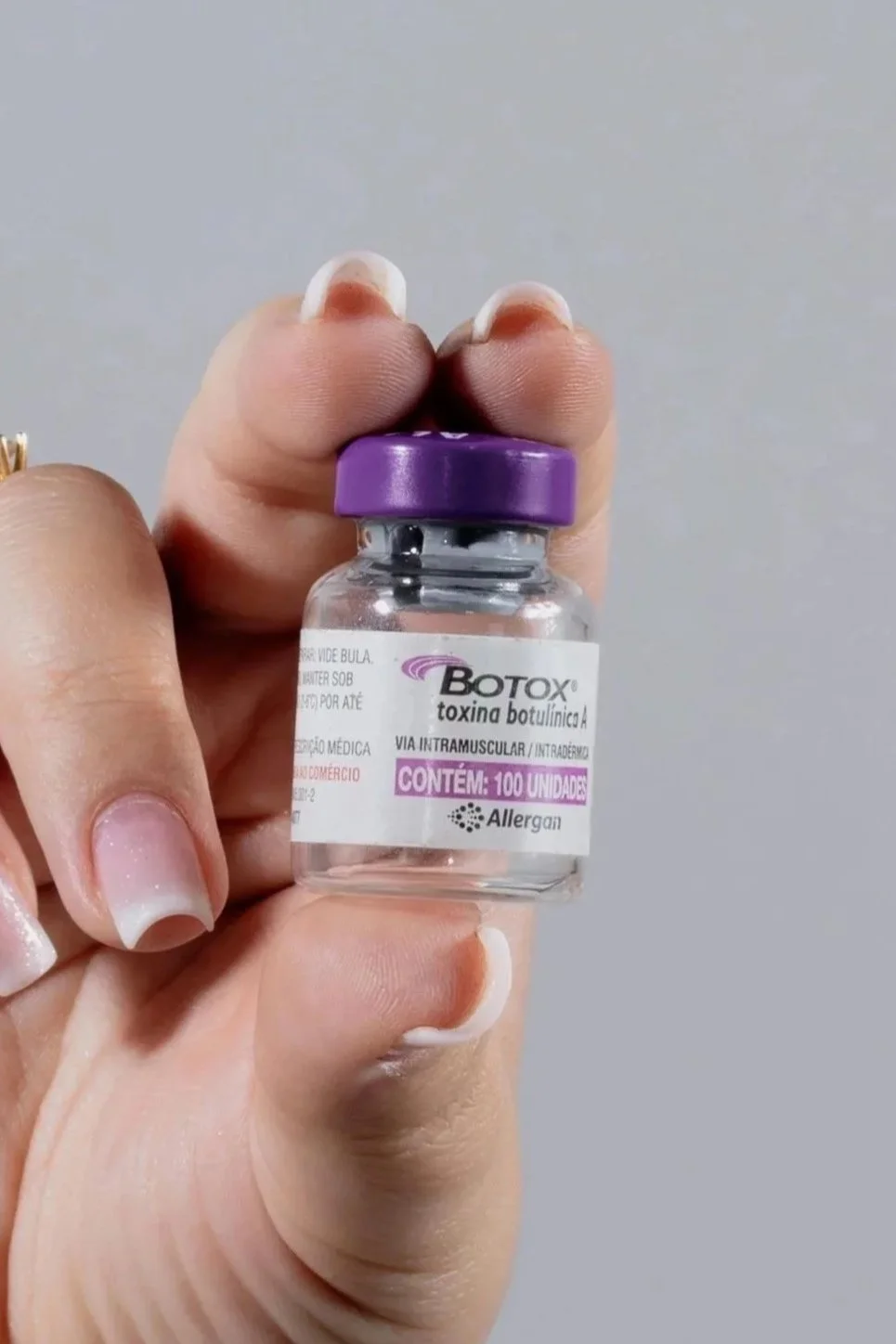 A hand holding a small vial labeled 'Botox toxin A' with a purple cap, containing 100 units, used for intramuscular injections.