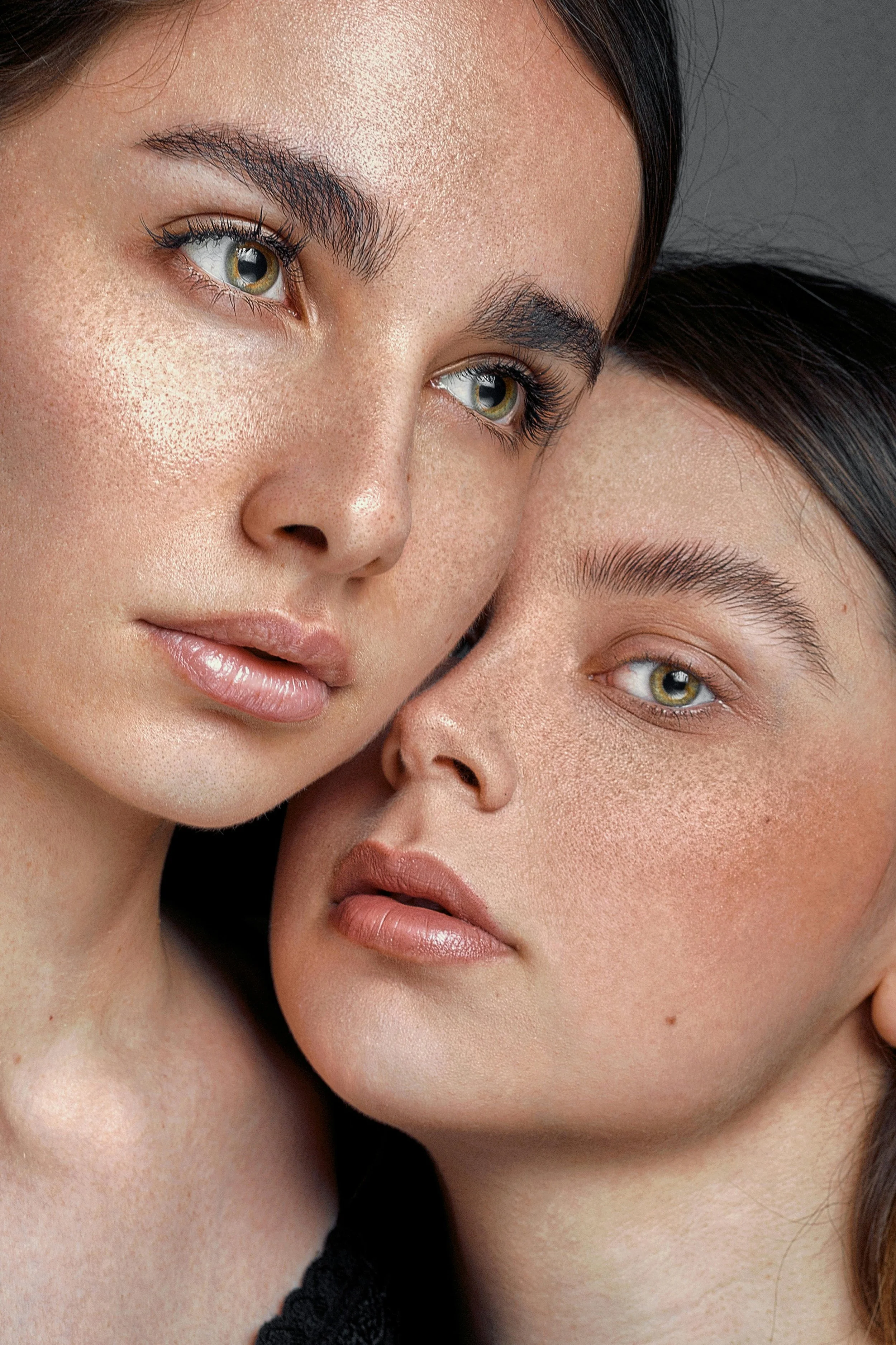 Close-up of two women with clear, fair skin, freckles, and minimal makeup, their faces touching, looking at the camera.