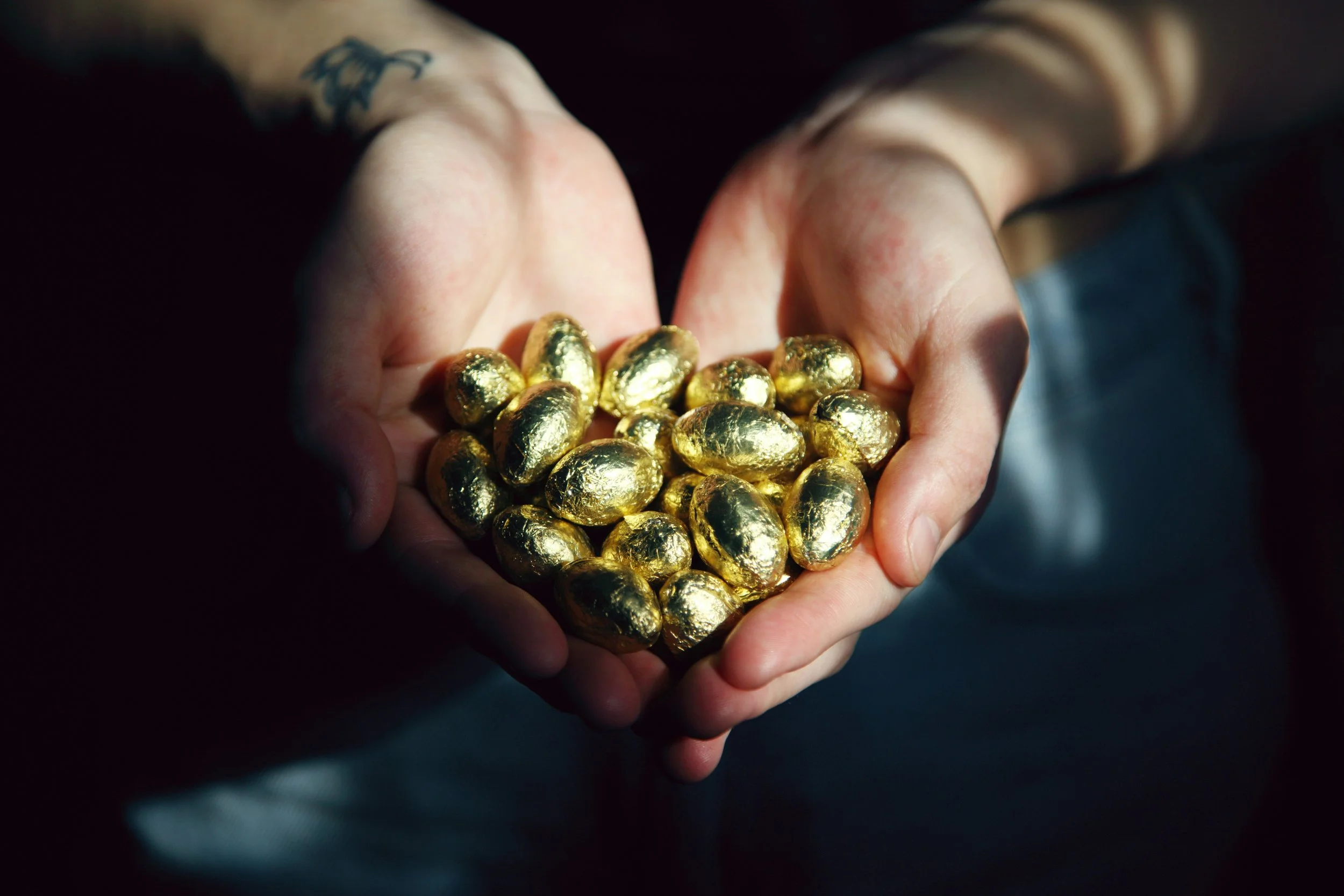 Hands holding a pile of shiny gold-wrapped chocolates in the shape of a heart.