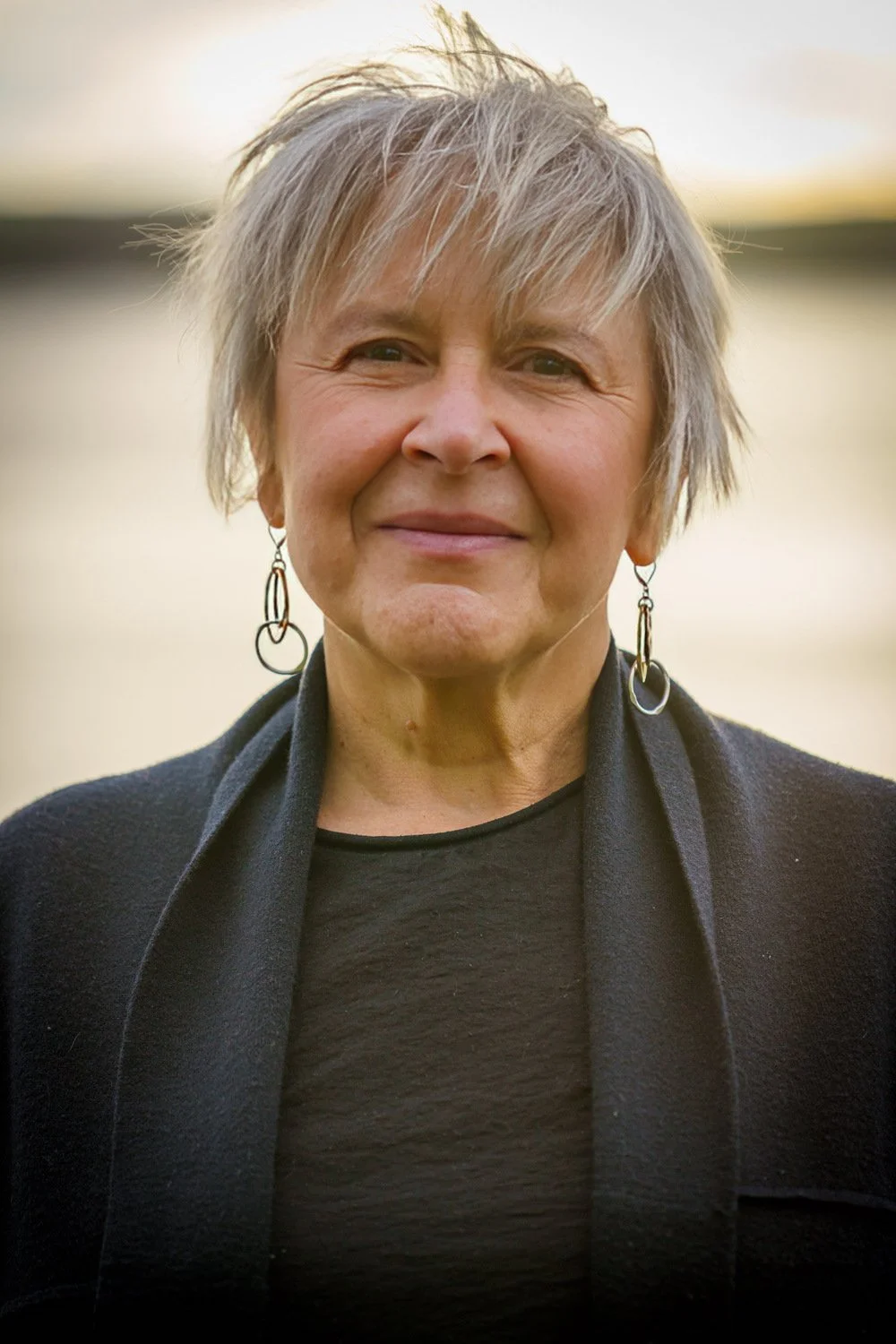 A senior woman with short, gray hair, smiling outdoors at sunset, wearing earrings, a black top, and a black jacket.
