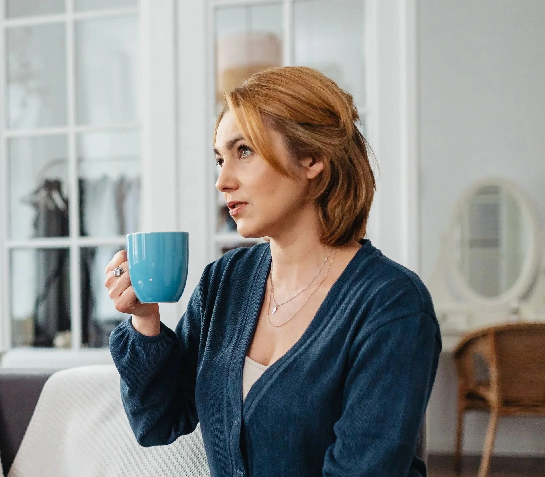 Woman in a blue cardigan sitting on a sofa, holding a blue mug and looking to the side.