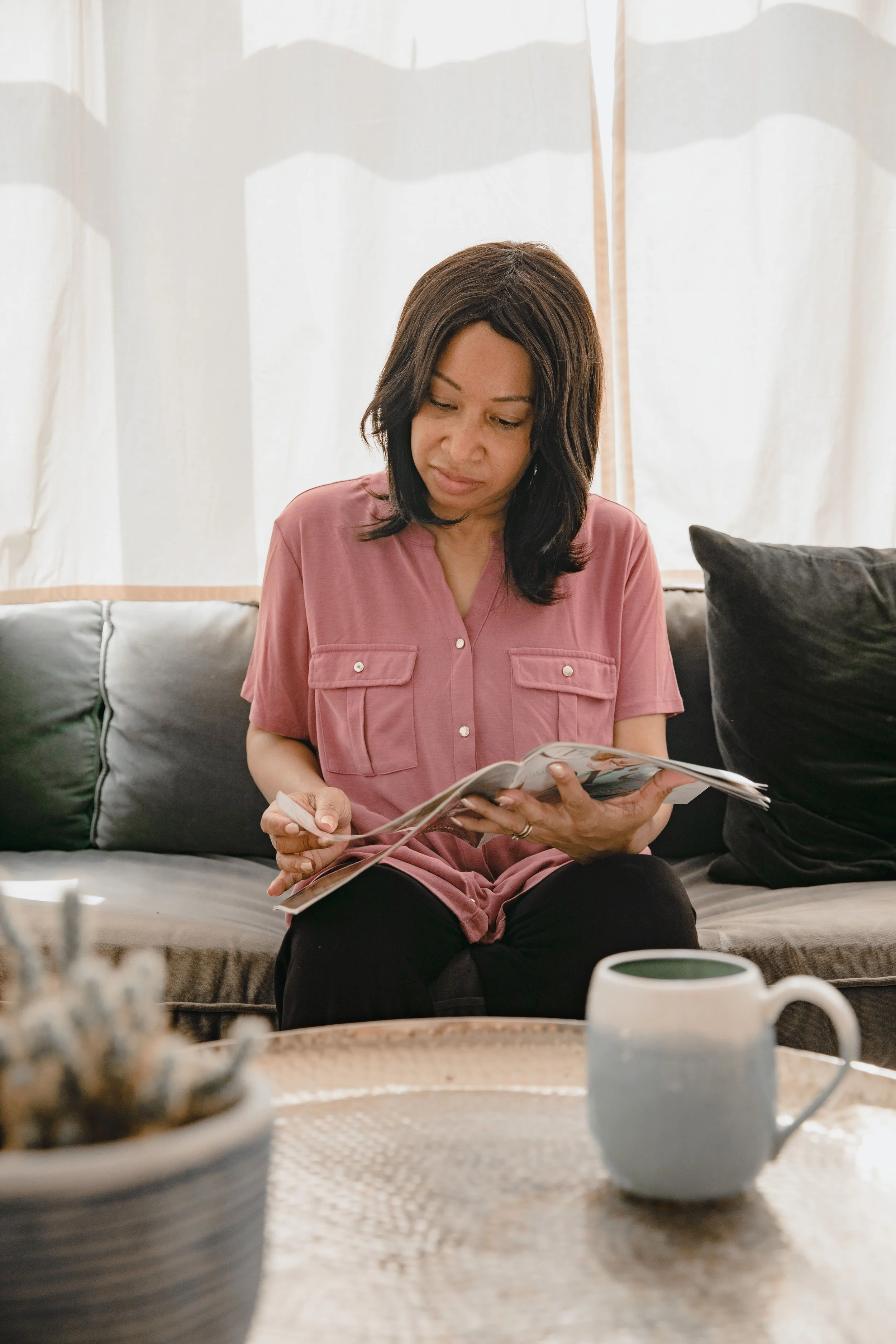 Woman in a pink shirt reading a magazine while seated on a sofa at home.