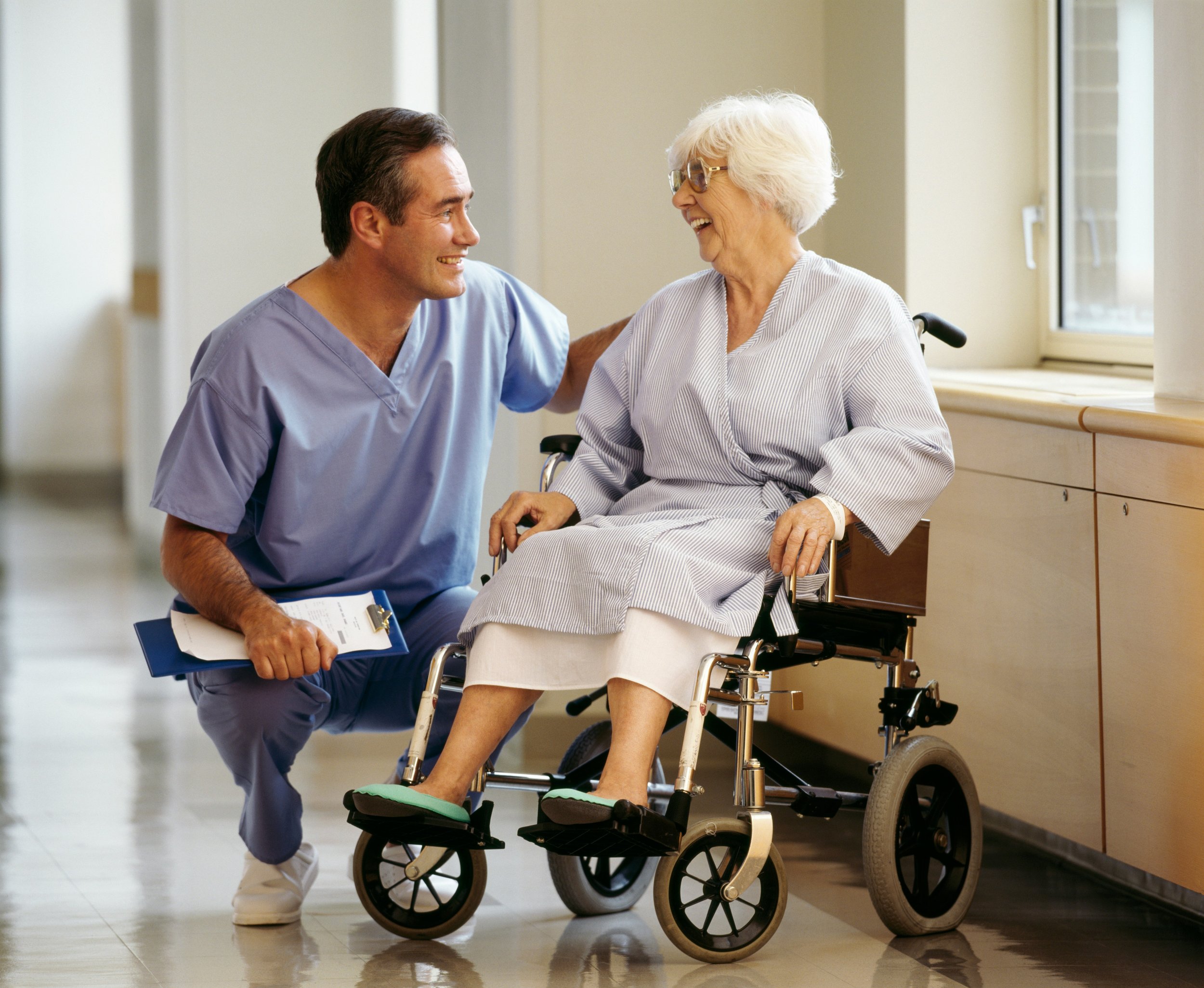 Smiling healthcare professional in scrubs kneeling beside an elderly woman in a wheelchair, inside a hospital setting.