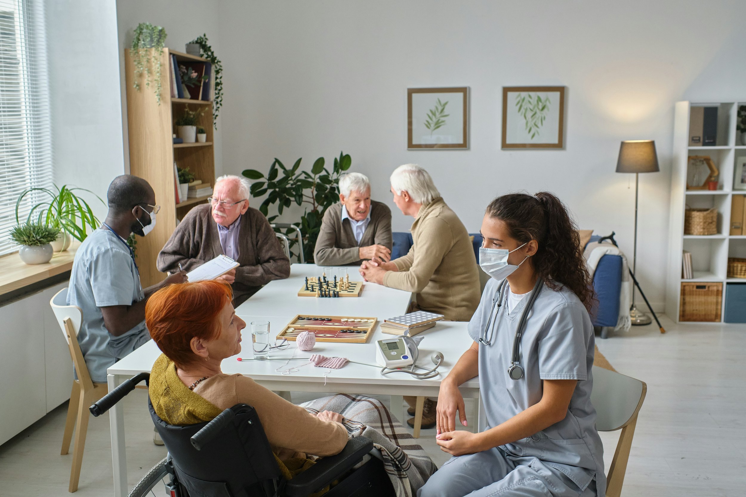 Caregivers interacting with seniors in a communal living space, engaging in conversations and activities like chess, with medical equipment visible nearby.
