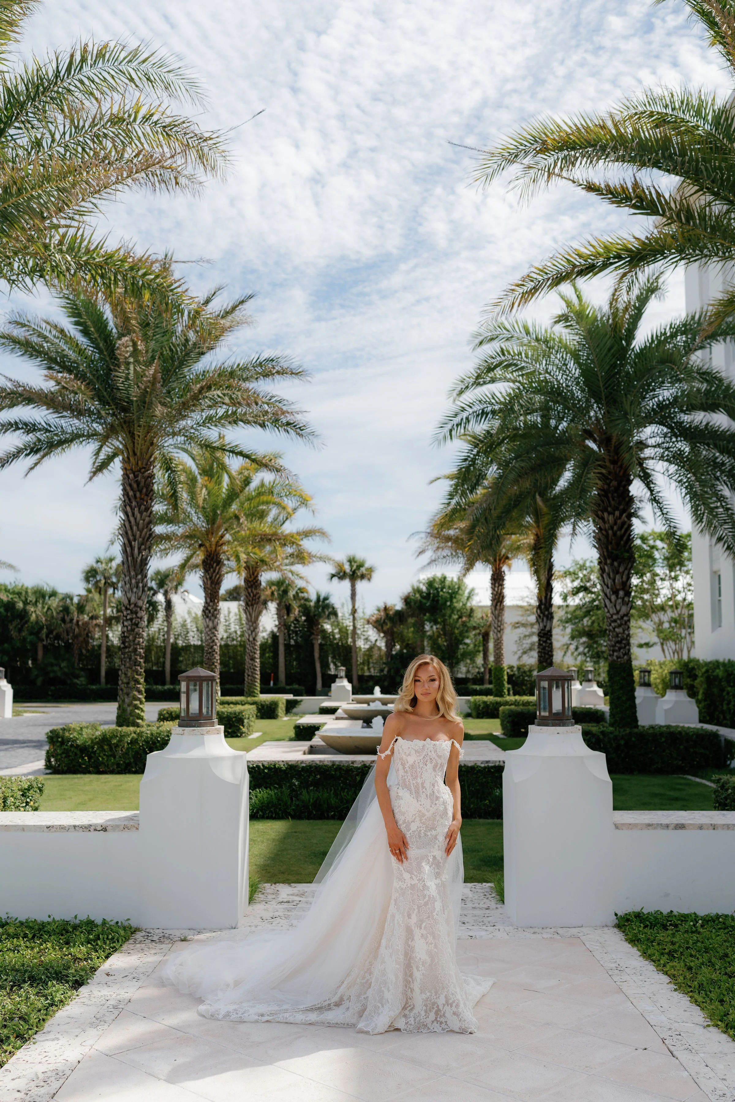 Full-length portrait of a luxury bride in an elegant embellished wedding dress with a long train, standing in a manicured palm tree garden walk planned by Carolyn Touchette Events in 30A.