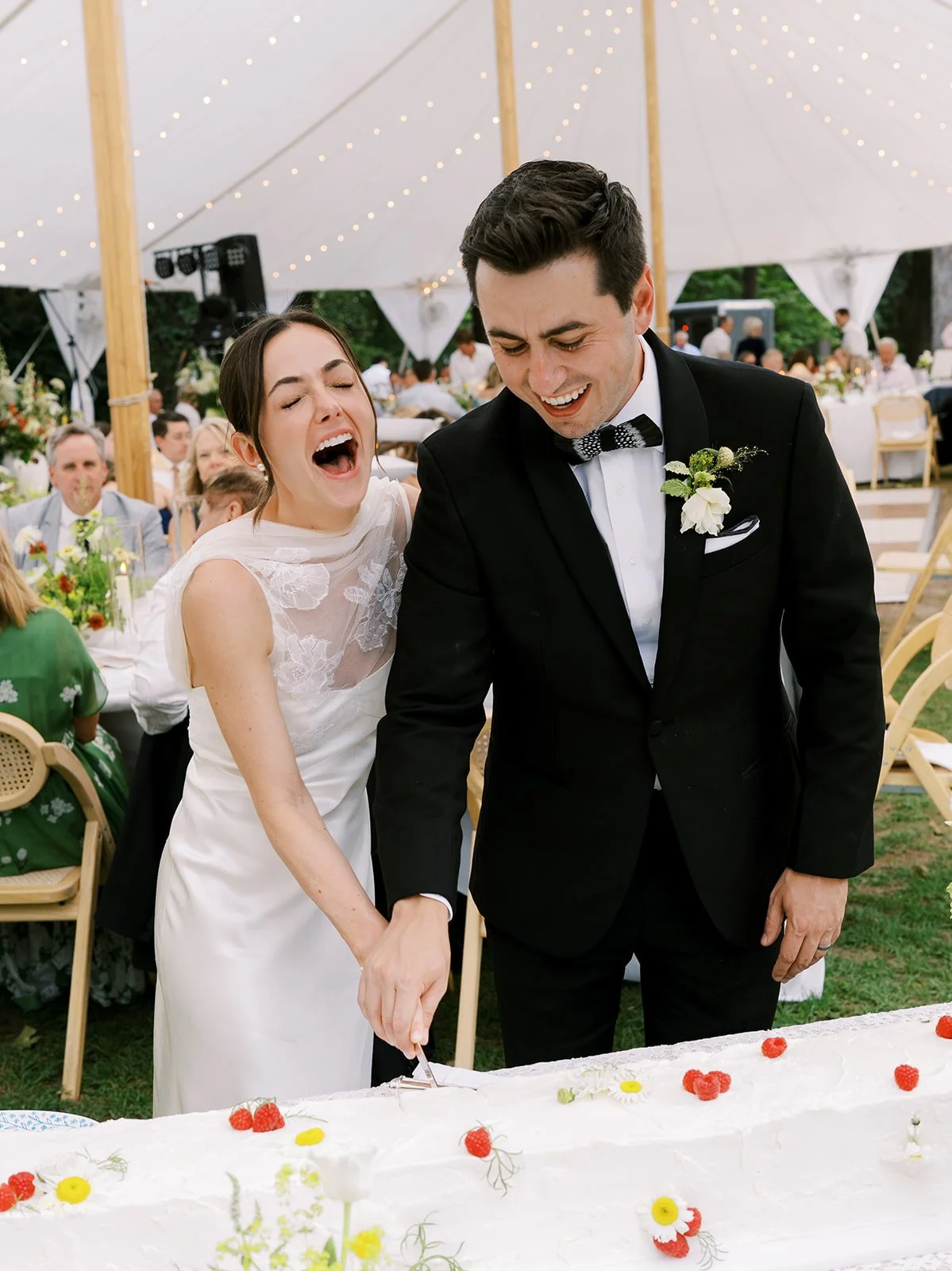 Joyful bride and groom laughing while cutting a large rectangular wedding cake topped with fresh berries and daisies under a tented reception, coordinated by Carolyn Touchette Events.
