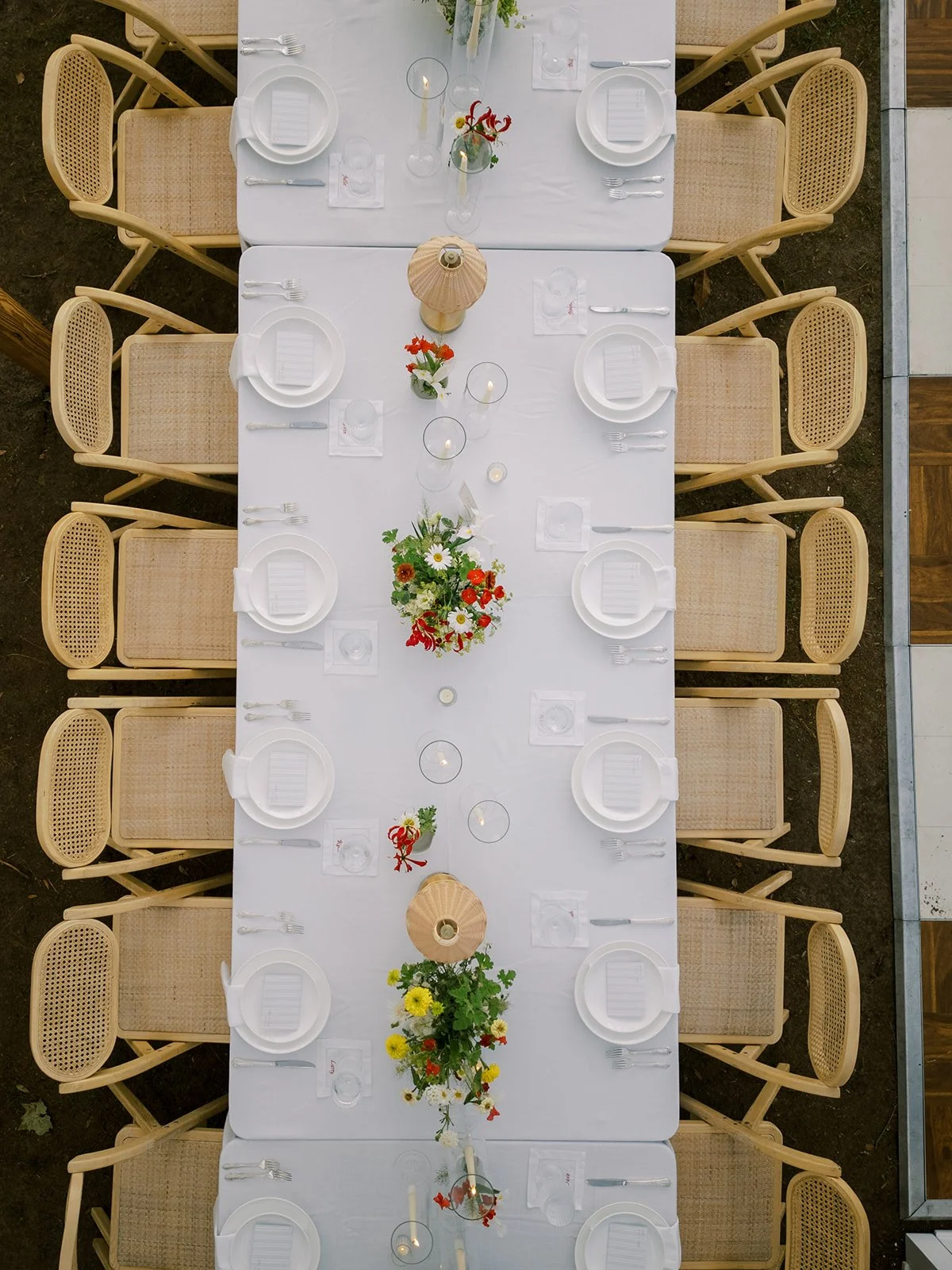 Direct overhead aerial view of a wedding reception dining table set with white china, woven cane chairs, floral arrangements, and warm table lamps, by Carolyn Touchette Events.