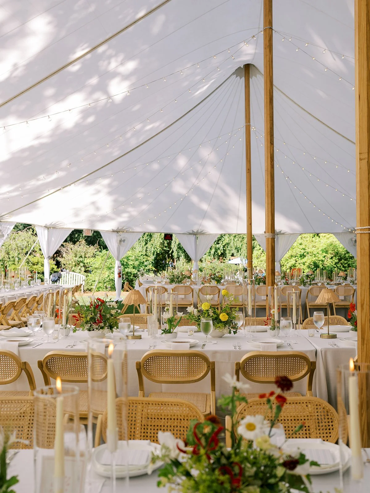 Interior view of a sailcloth tented wedding reception featuring long dining tables, woven rattan chairs, cordless table lamps, and floral centerpieces, styled by Carolyn Touchette Events.