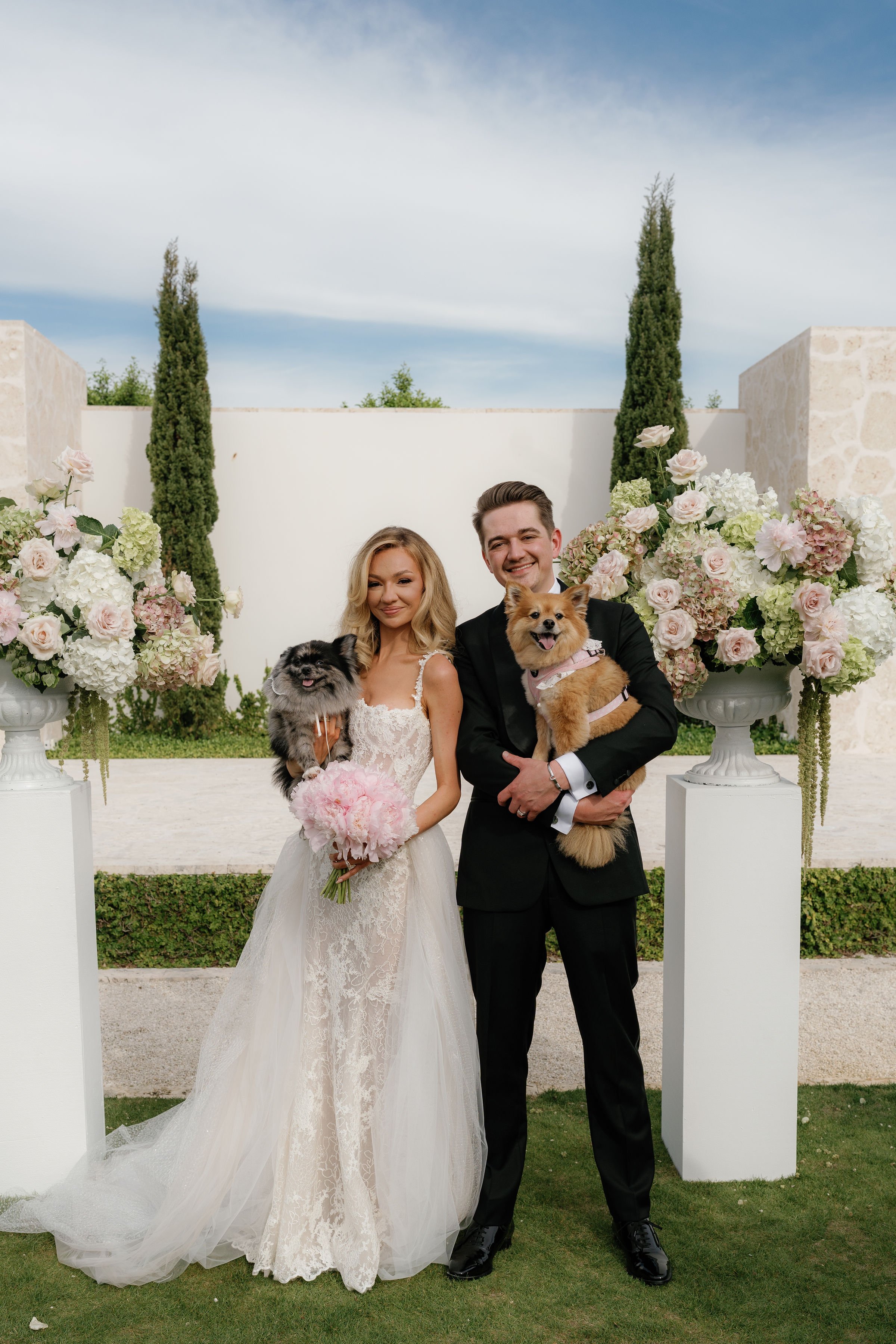 Smiling bride and groom holding their two dogs at a pet-friendly luxury outdoor wedding ceremony altar with pastel floral pedestals, planned by Carolyn Touchette Events.
