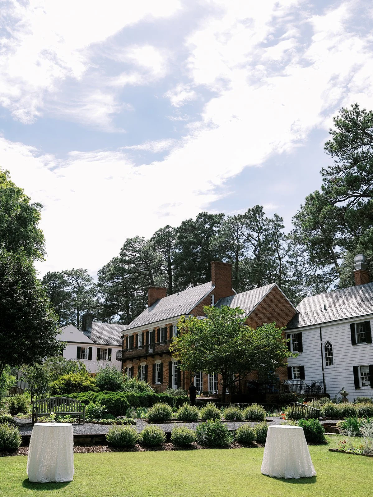 Exterior view of a historic brick estate wedding venue in Pinehurst, featuring outdoor cocktail hour high-top tables on a landscaped garden lawn, coordinated by Carolyn Touchette Events.