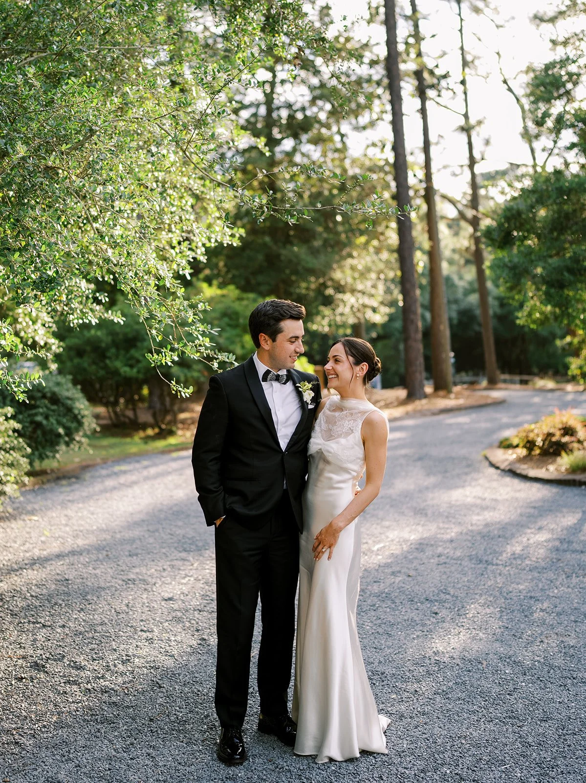 Romantic portrait of a bride in a silk gown and groom in a classic tuxedo walking down a tree-lined gravel path, planned by Carolyn Touchette Events.