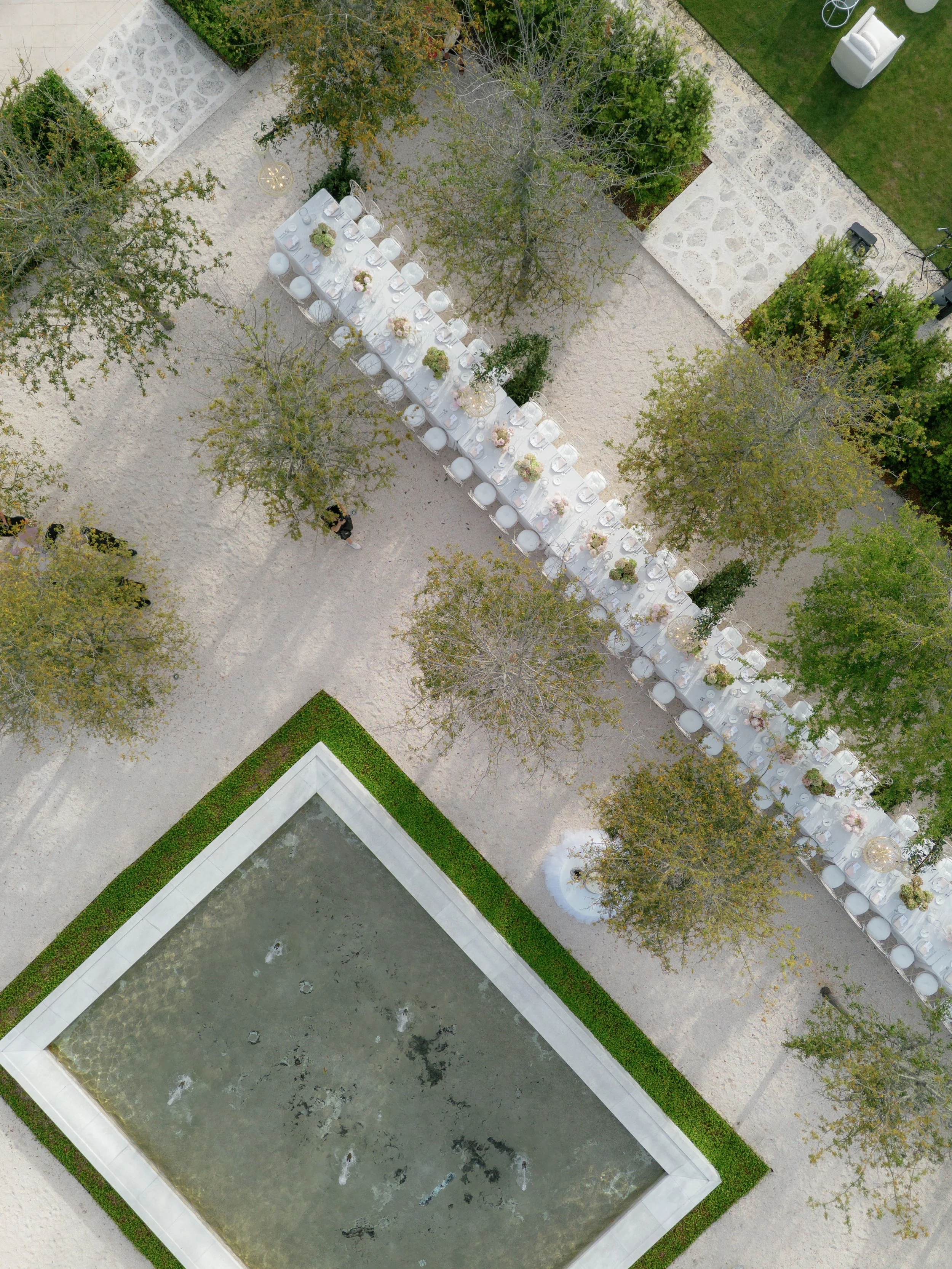 Direct overhead aerial view of a long white wedding reception dining table on a gravel courtyard with a square reflection pool, showing a complete outdoor event layout by Carolyn Touchette Events.