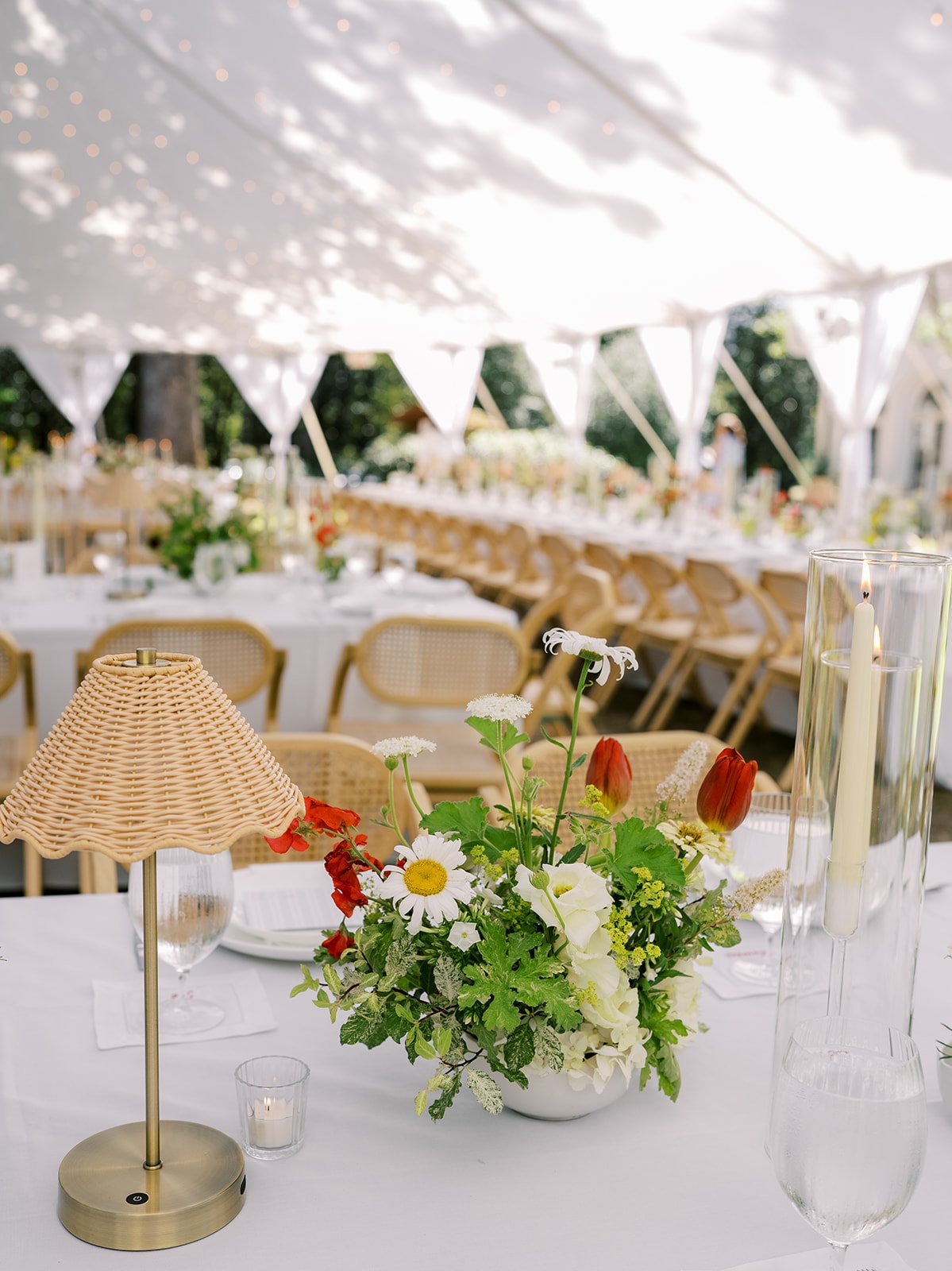 Organic wedding centerpiece with ranunculus, tulips, and delphinium on a long reception table under a sailcloth tent with light dappling, designed by Carolyn Touchette Events in New England.