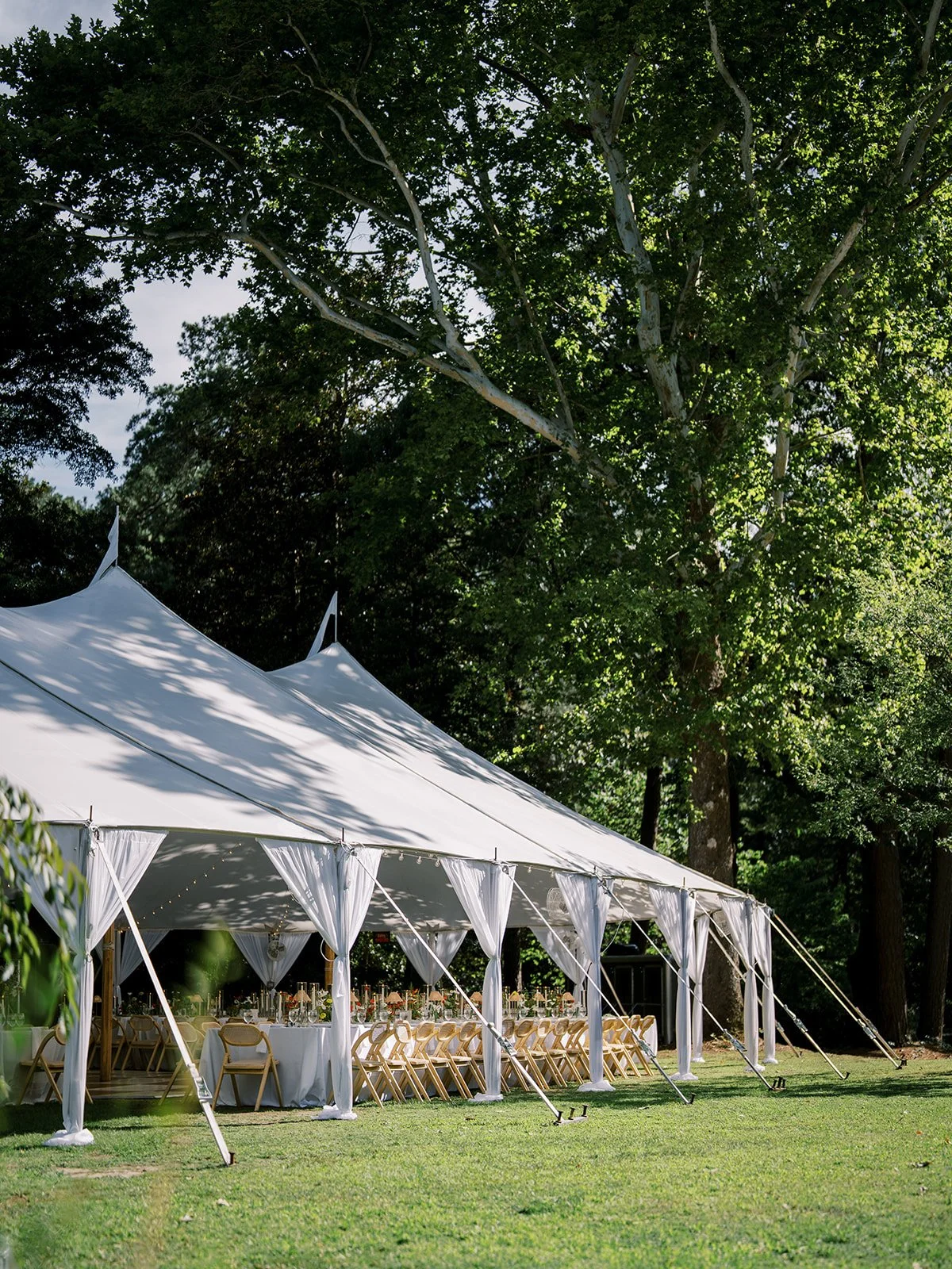 Lush outdoor setting for a luxury tented wedding reception with draped white fabric, string lights, and long dining tables on a manicured lawn by Carolyn Touchette Events.