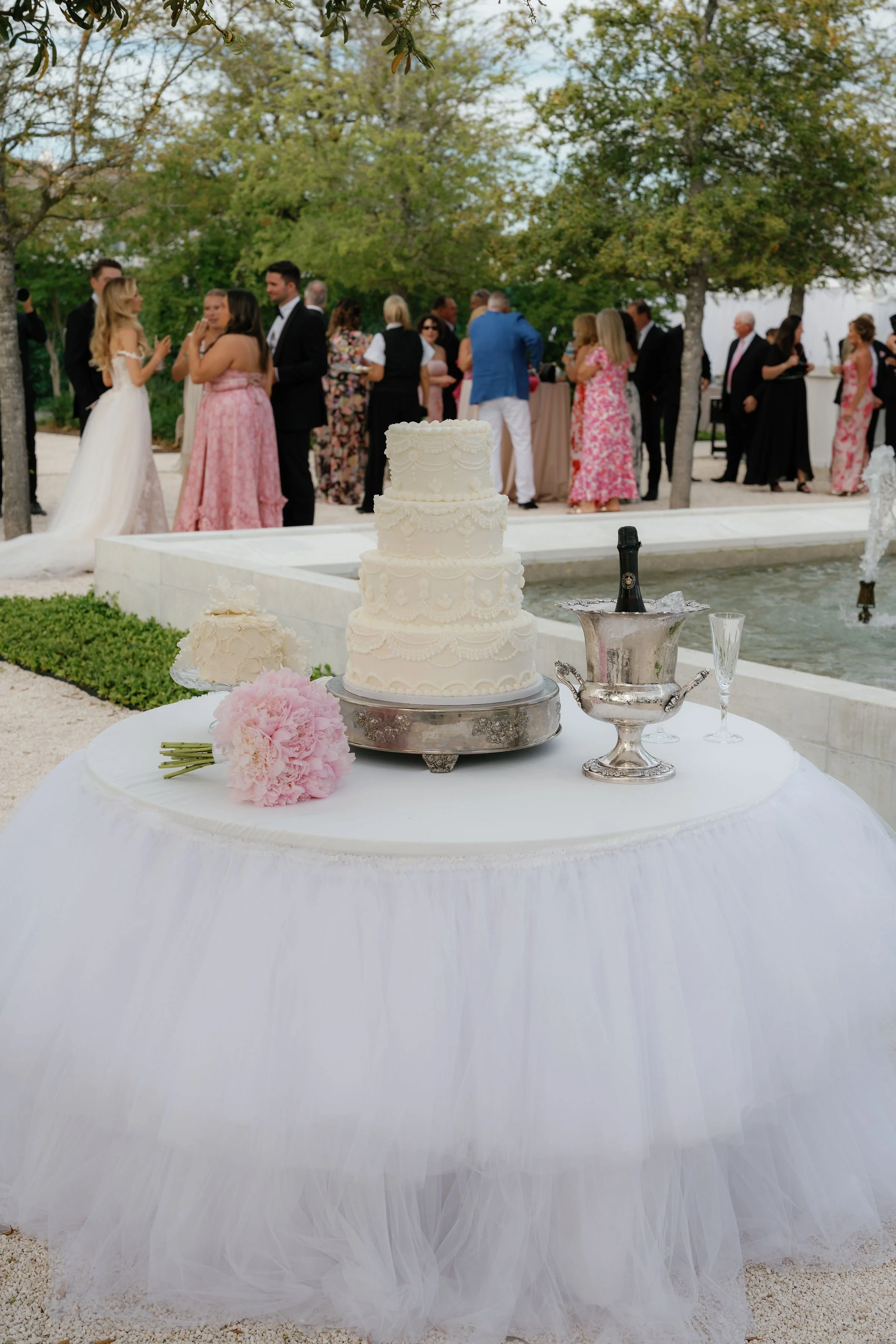 Four-tier classic white wedding cake and silver champagne bucket displayed on a custom white tulle table next to a formal garden fountain, planned by Carolyn Touchette Events in 30A, Florida.