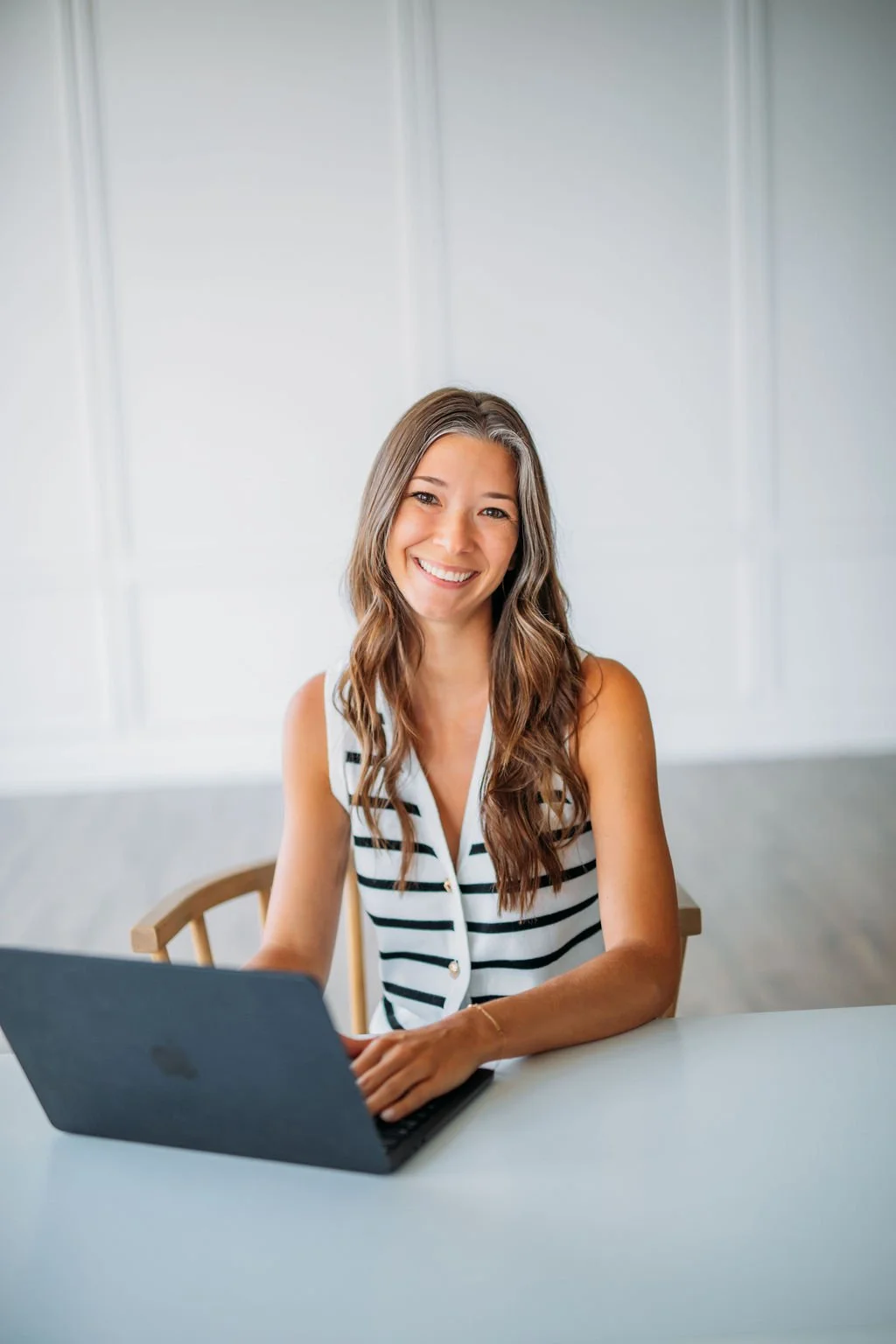A woman with long wavy brown hair smiling and sitting at a table with a laptop in front of her.