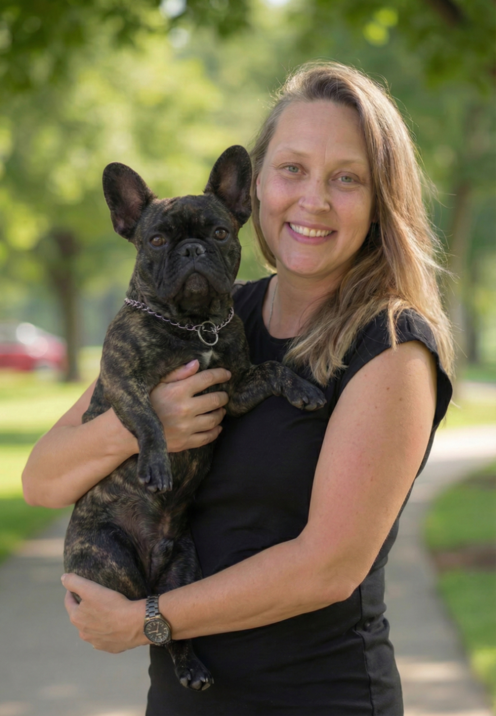 A woman with long light brown hair smiling while holding a brindle French Bulldog puppy outdoors on a sunny day.