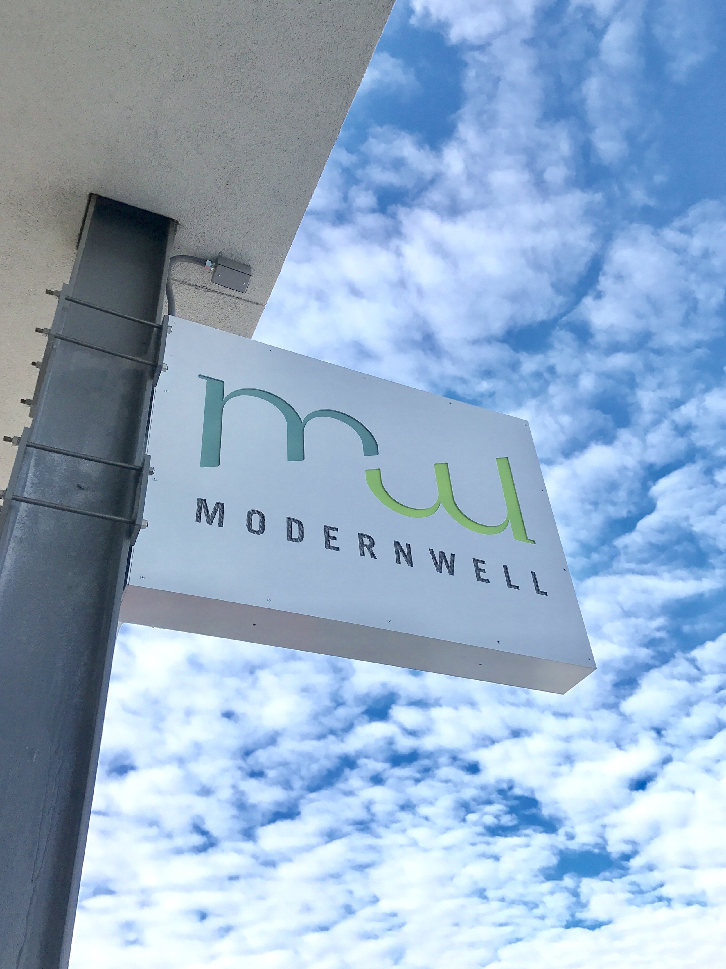 Exterior sign for Modernwell with blue sky and clouds in the background.