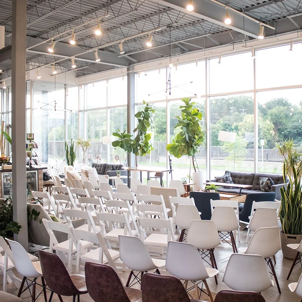 Interior of a brightly lit modern cafe or event space with white chairs arranged in rows, large glass windows with views of greenery, and indoor potted plants.