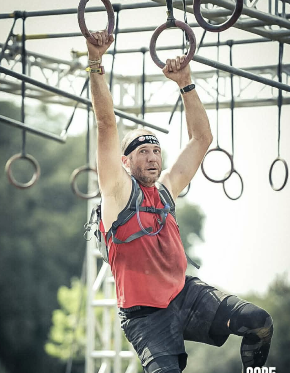 Man participating in an obstacle course, hanging from gymnastic rings with trees and equipment in the background.