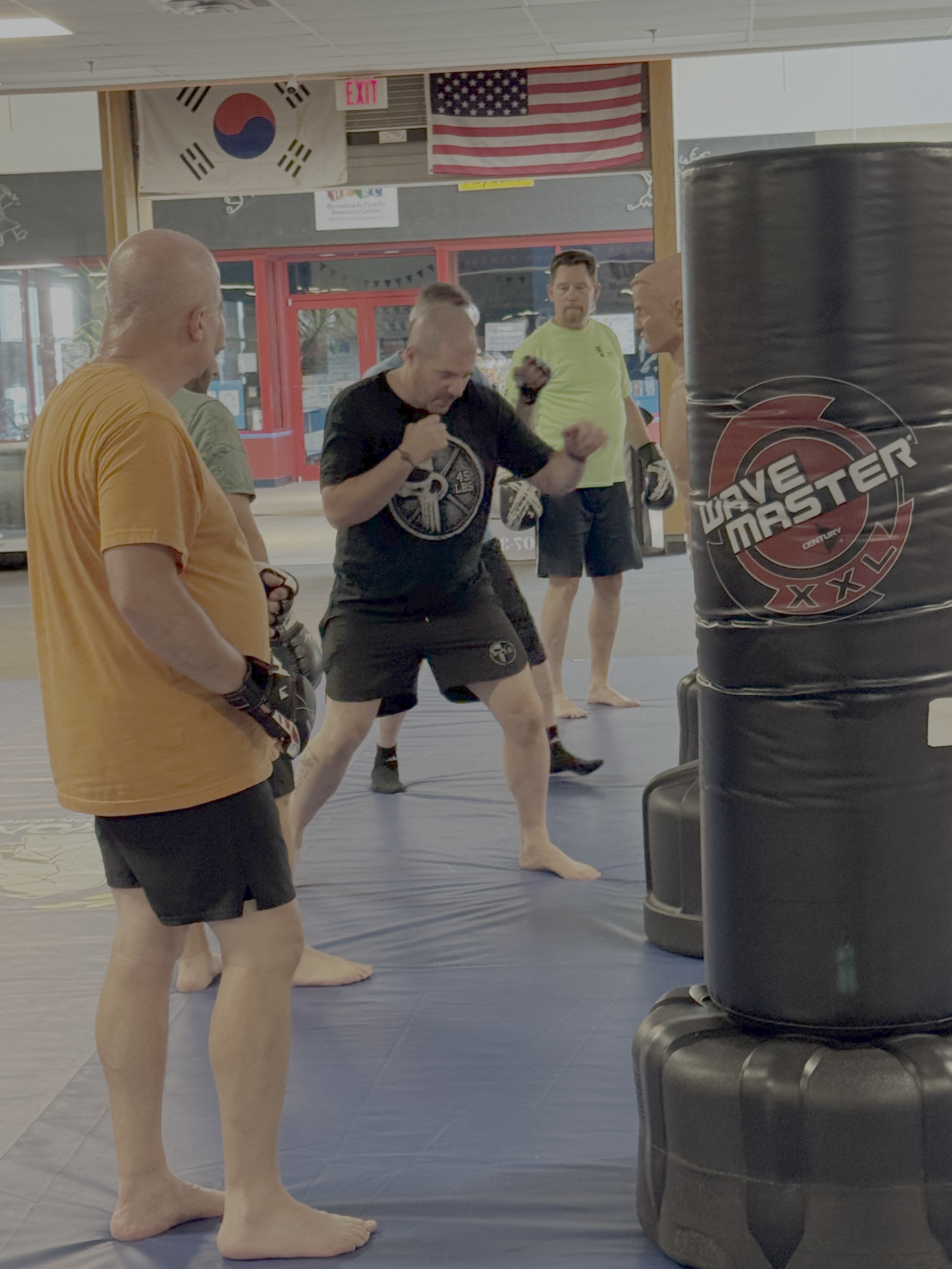 A group of people practicing martial arts or boxing in a gym, with a punching bag in the foreground, and flags of South Korea and the United States hanging on the wall in the background.
