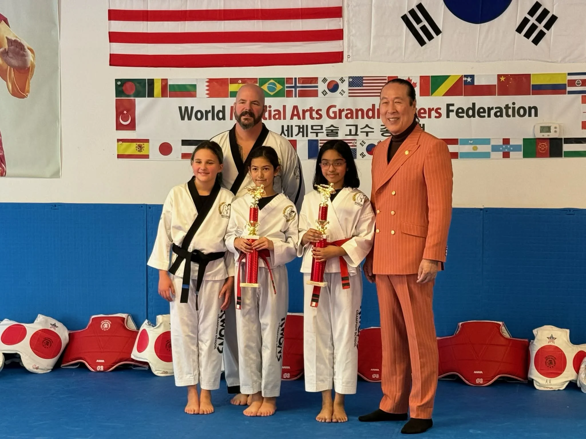 Three young girls in martial arts uniforms holding trophies, standing with two men in a martial arts dojo, with a banner and flags of various countries in the background.