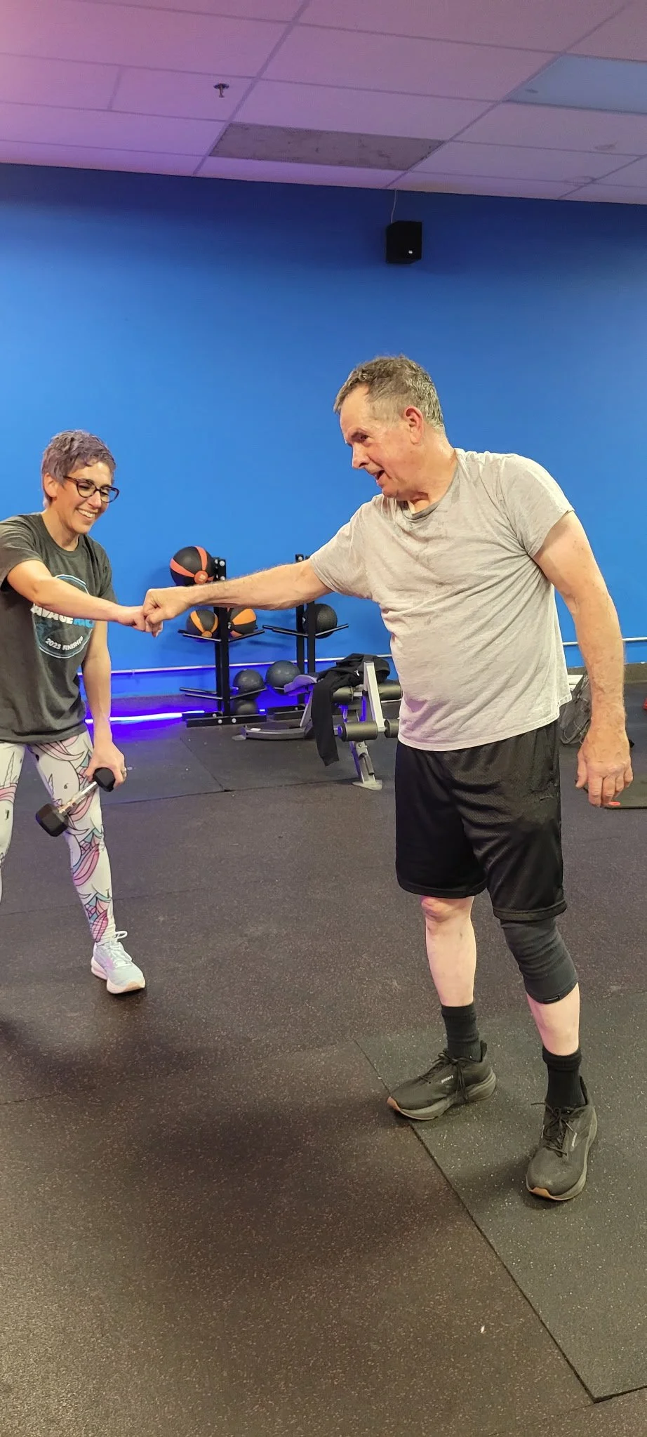 A woman and a man are in a gym, doing a fist bump. The woman is holding a dumbbell, and both are smiling. The gym has various exercise equipment in the background, with bright lighting and a blue wall.