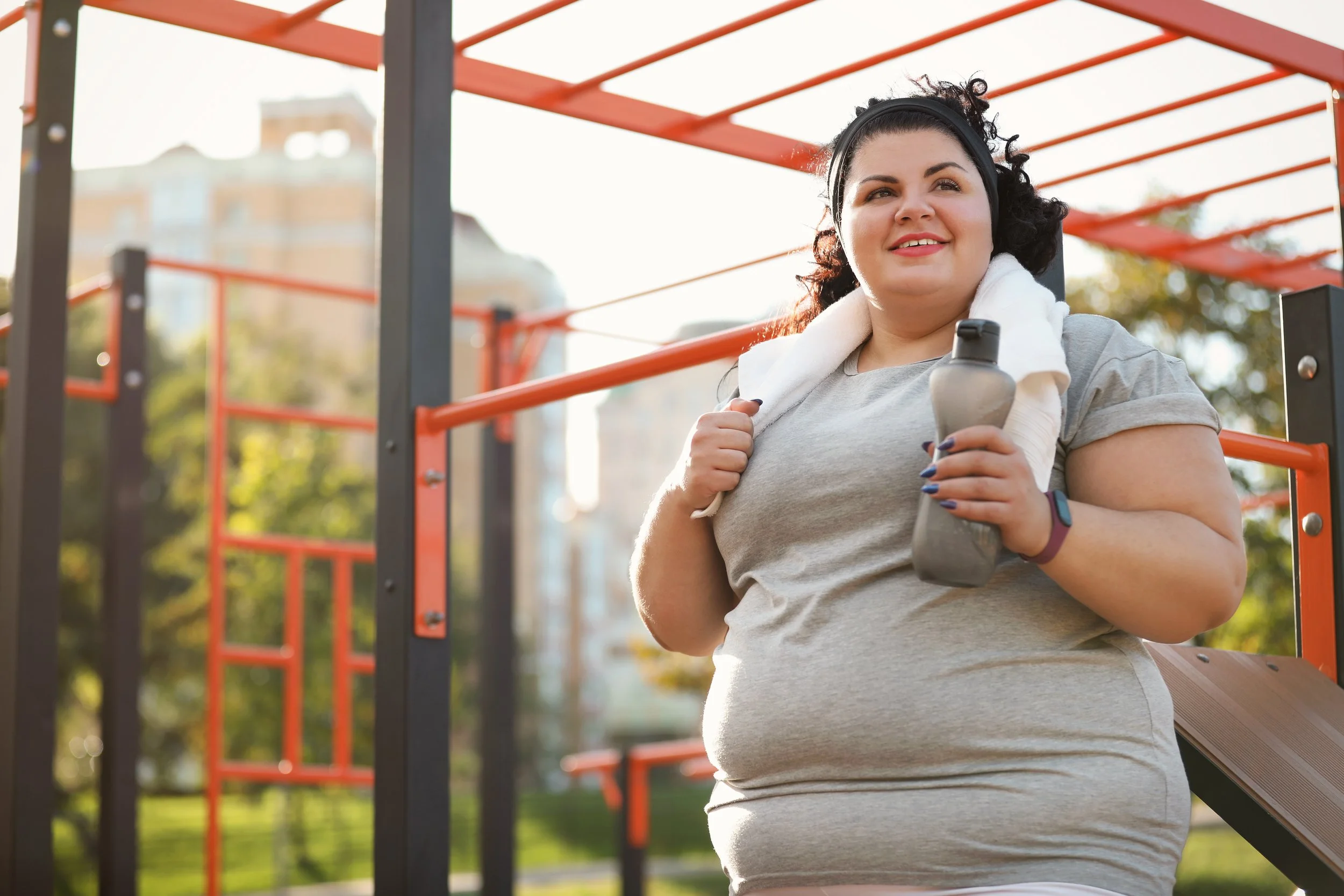 A woman at a playground holding a water bottle, with black curly hair, wearing a gray t-shirt and a towel around her neck, enjoying sunny outdoor weather.