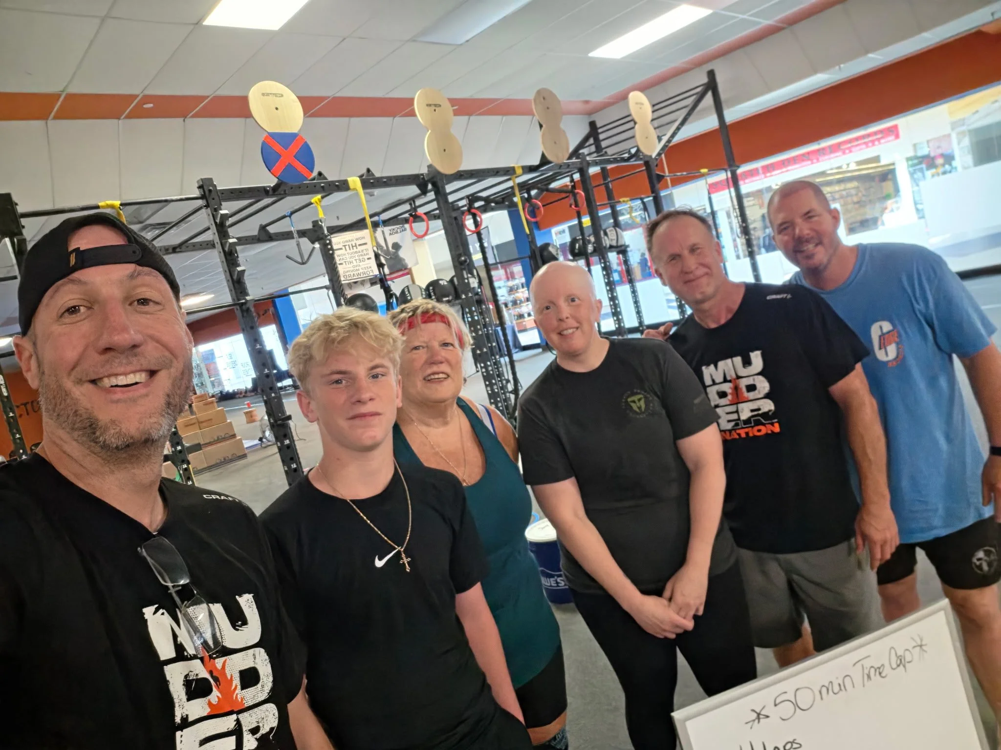 Group of people smiling in a gym, standing in front of workout equipment and a whiteboard with workout instructions.