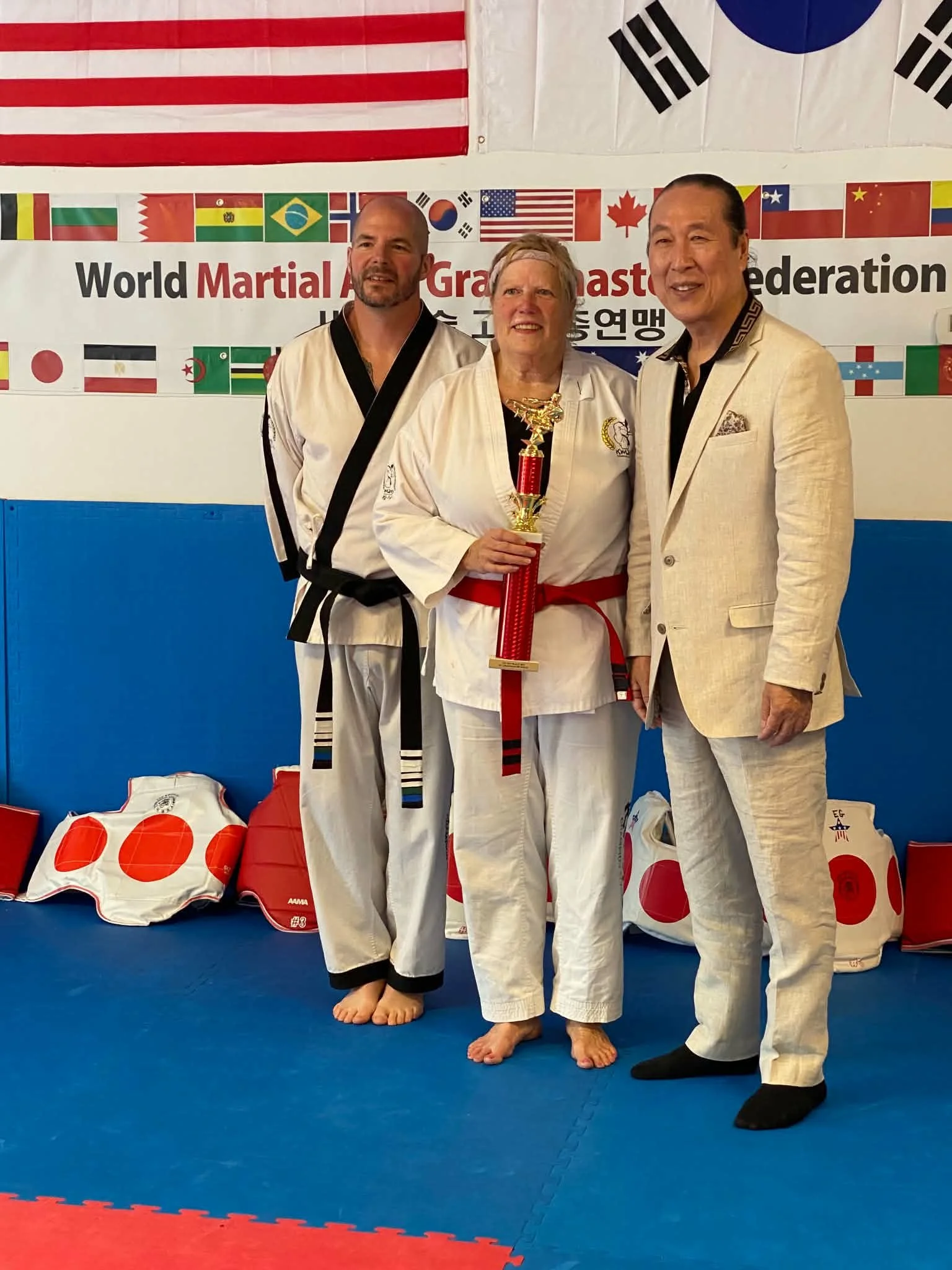 Three martial artists in traditional uniforms standing together on a blue and red mat, holding a trophy, with national flags of the United States, South Korea, and other countries hanging behind them at a martial arts event.
