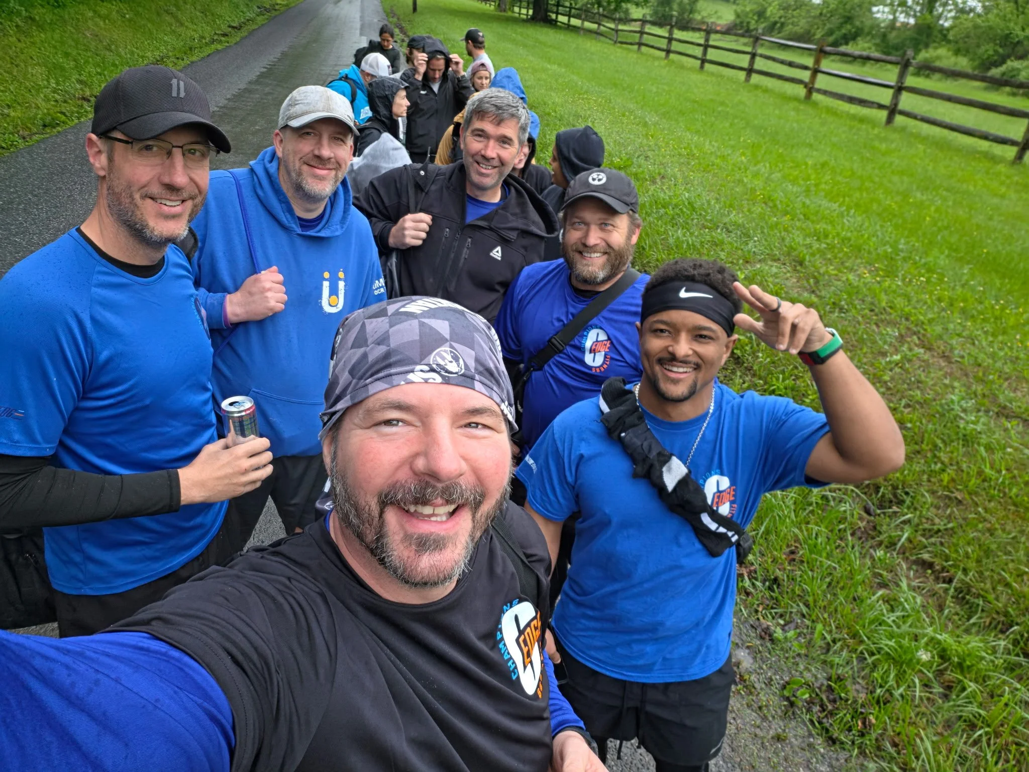 A group of people taking a selfie outdoors on a rainy day, with some wearing rain jackets and hats, standing on a grassy area with a wooden fence and trees in the background.