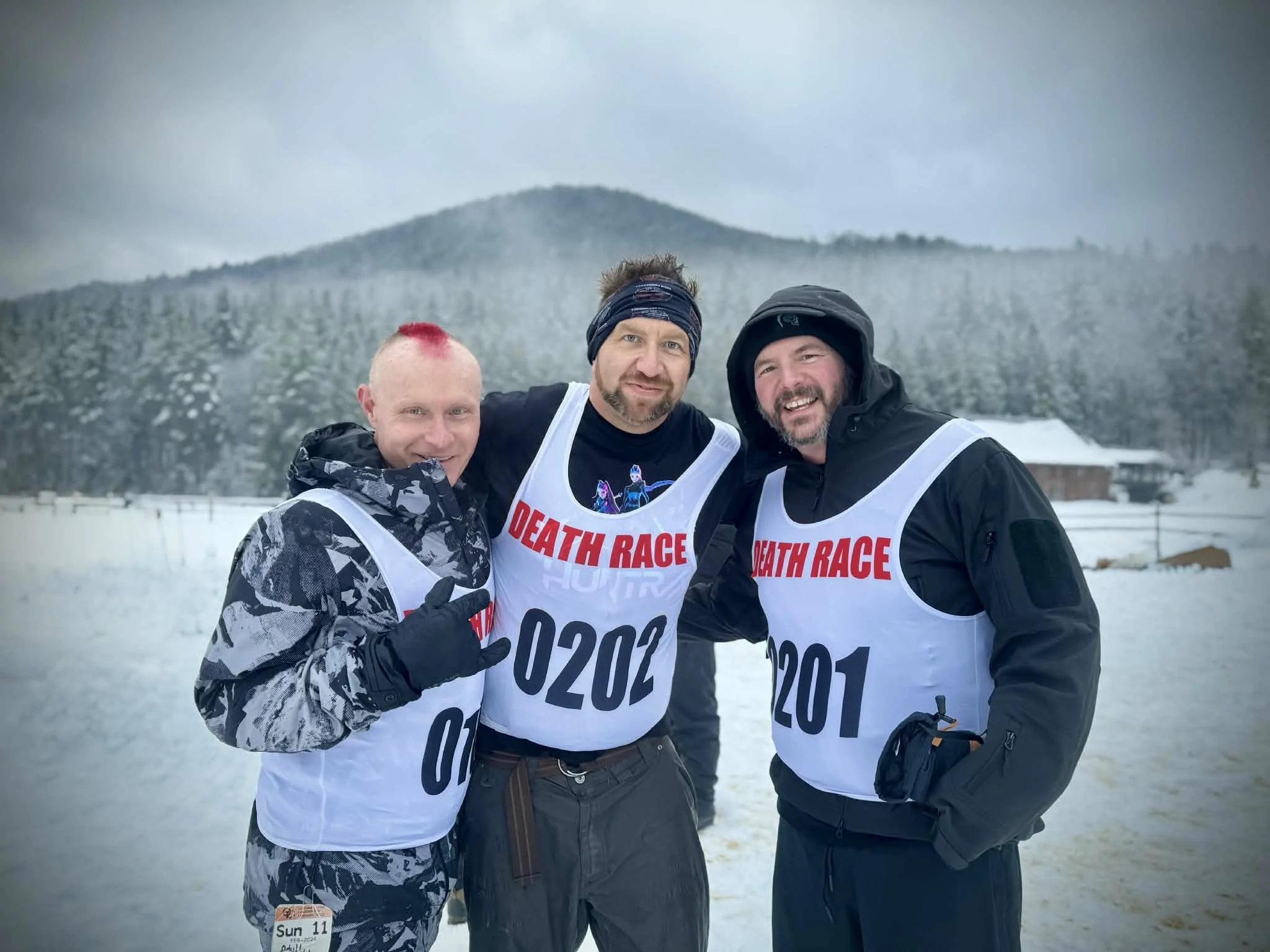 Three men standing outdoors in the snow, wearing athletic gear and race bibs, smiling for a photo during a snow race event, with mountains and cloudy sky in the background.