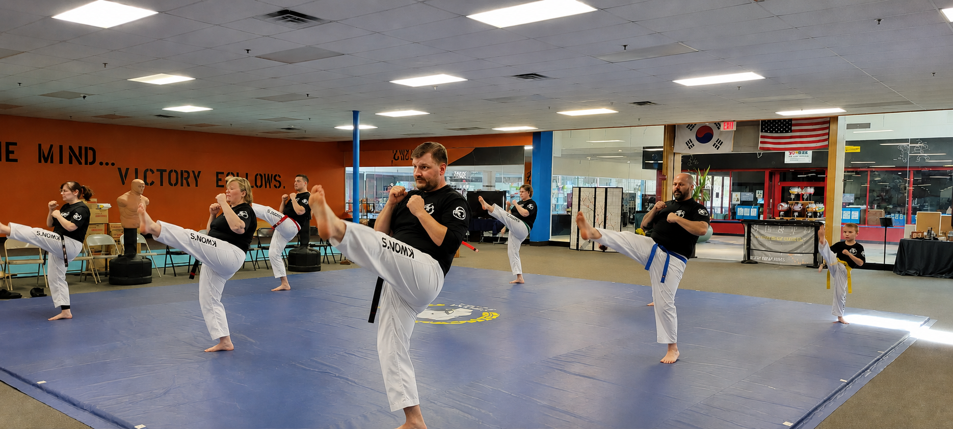 Martial arts class practicing kickboxing techniques on blue mats inside a gym, with instructors and students of various ages wearing black and white uniforms.