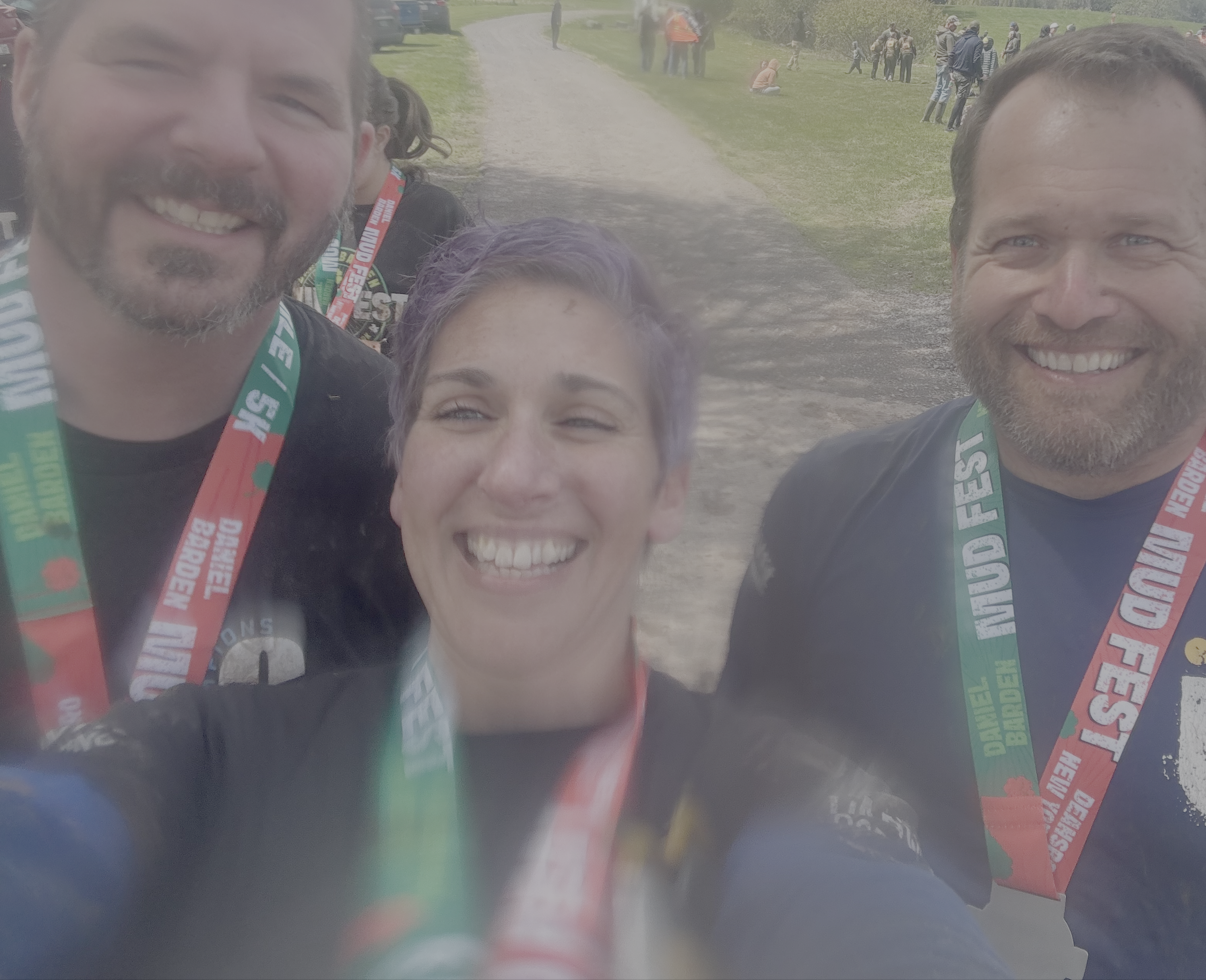 Close-up of three smiling people wearing medals and race bibs at an outdoor event, with a grassy area, dirt path, and a group of people in the background.