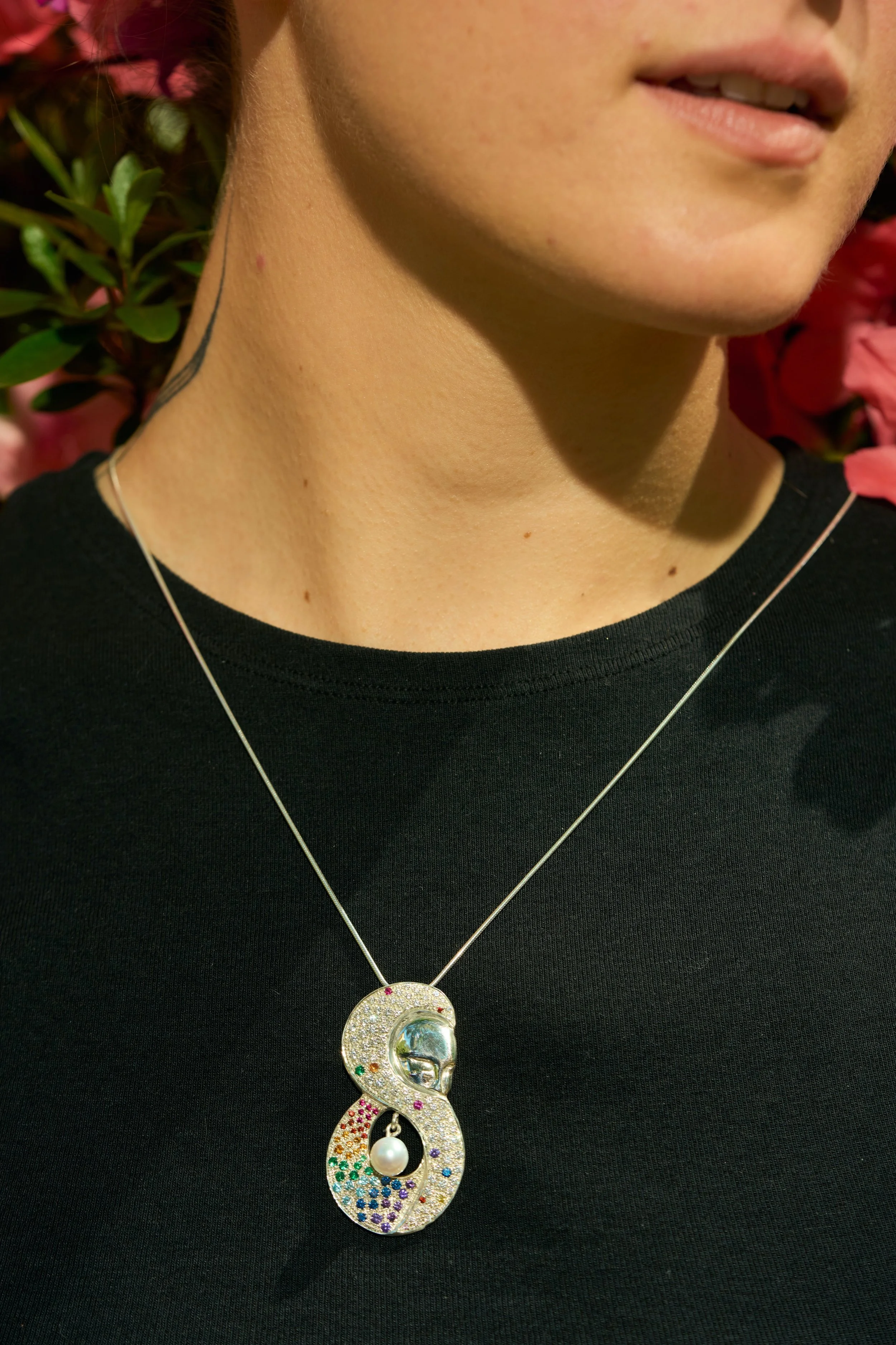 Close-up of a person's neck and lower face, showing part of a silver necklace, black shirt, and a decorative necklace with a large, colorful pendant featuring a pearl and rainbow rhinestones, with pink flowers in the background.
