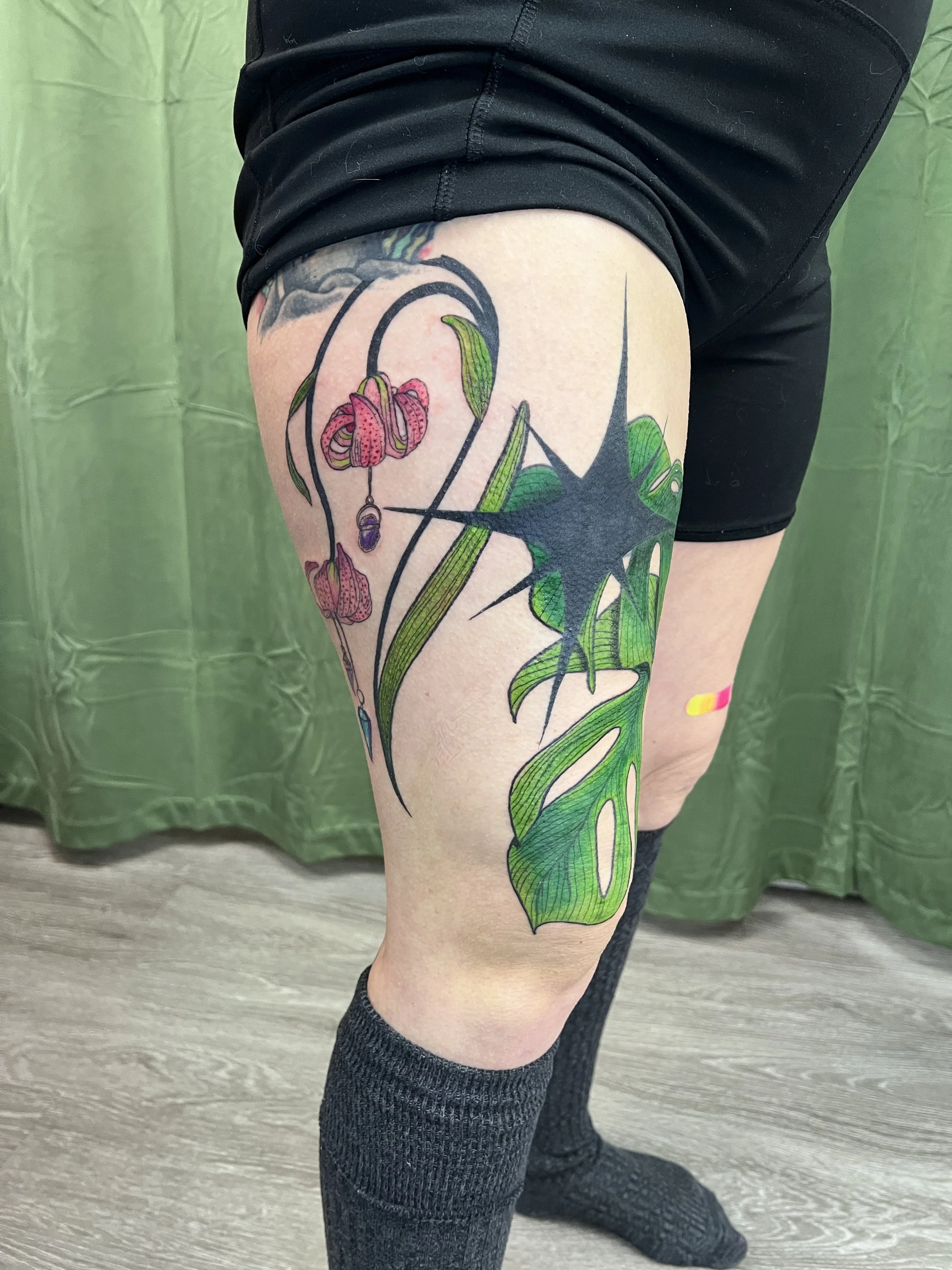 Close-up of a person's upper thigh and knee, revealing a colorful tattoo of pink flowers with green leaves and stems, with a black star and string of purple stones. The person is wearing black shorts and black knee-high socks, standing on a light-col