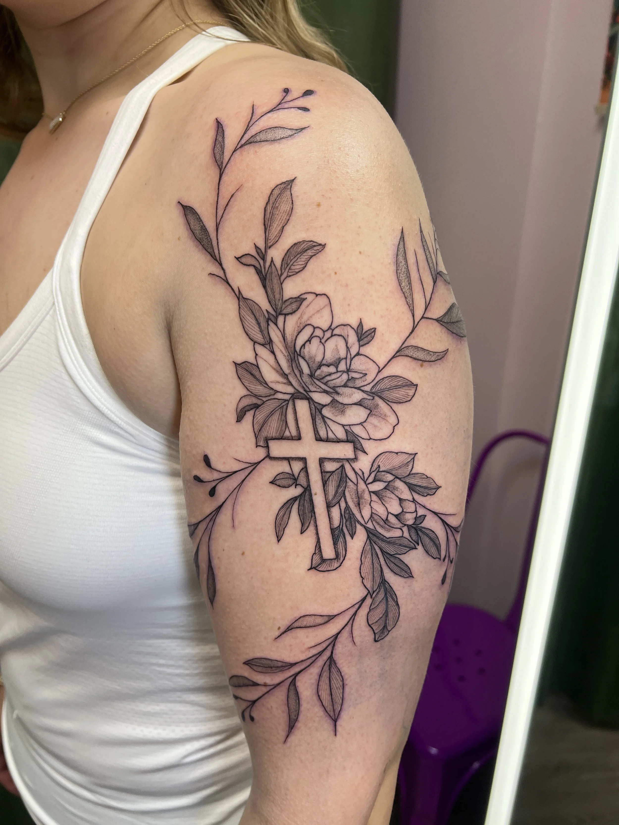 A person's upper arm with a black outline floral tattoo featuring leaves and flowers, and a cross incorporated into the design.
