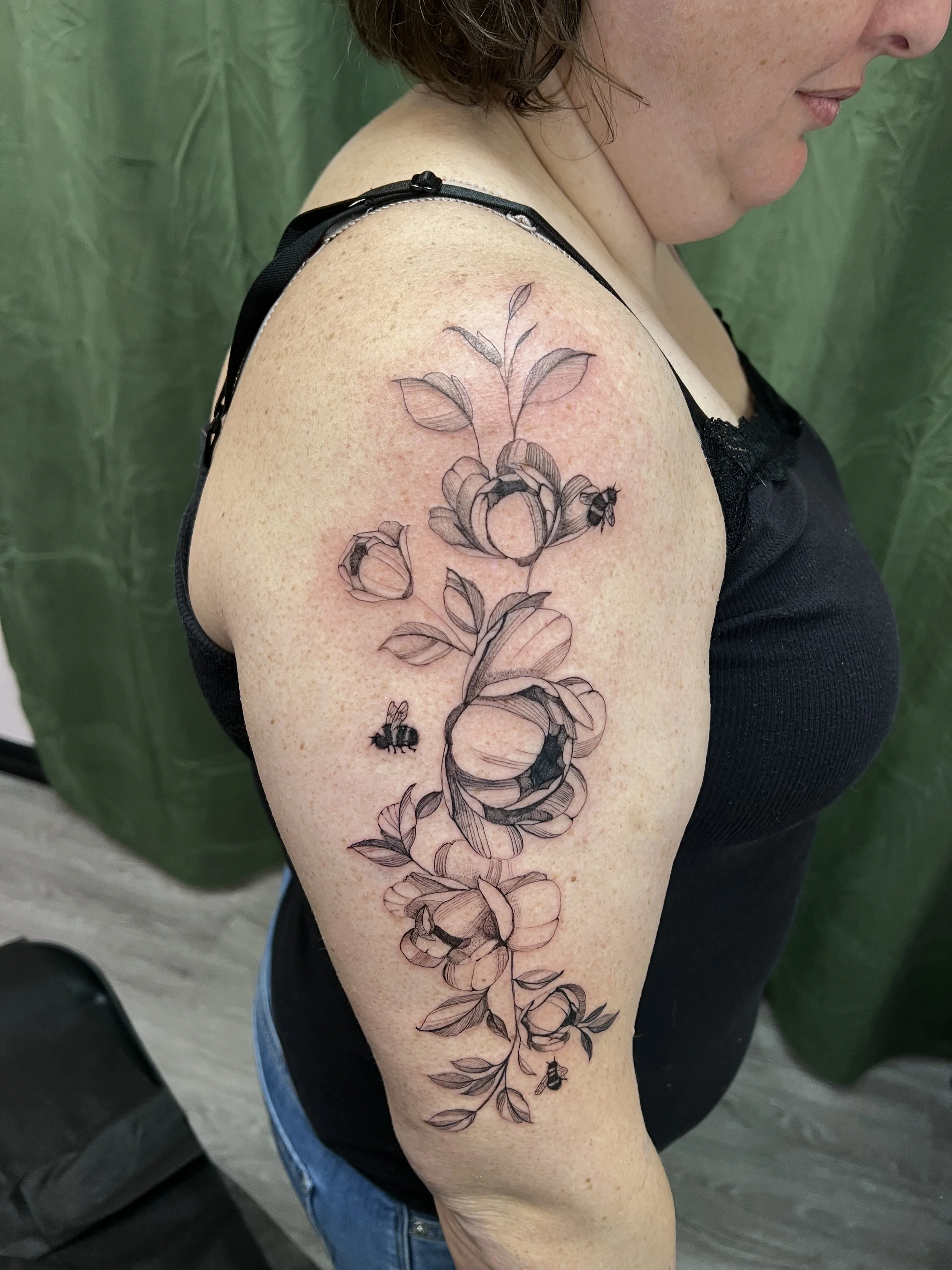 A woman with a black line drawing tattoo of a flower and bee on her upper arm, wearing a black sleeveless top, with a green curtain in the background.
