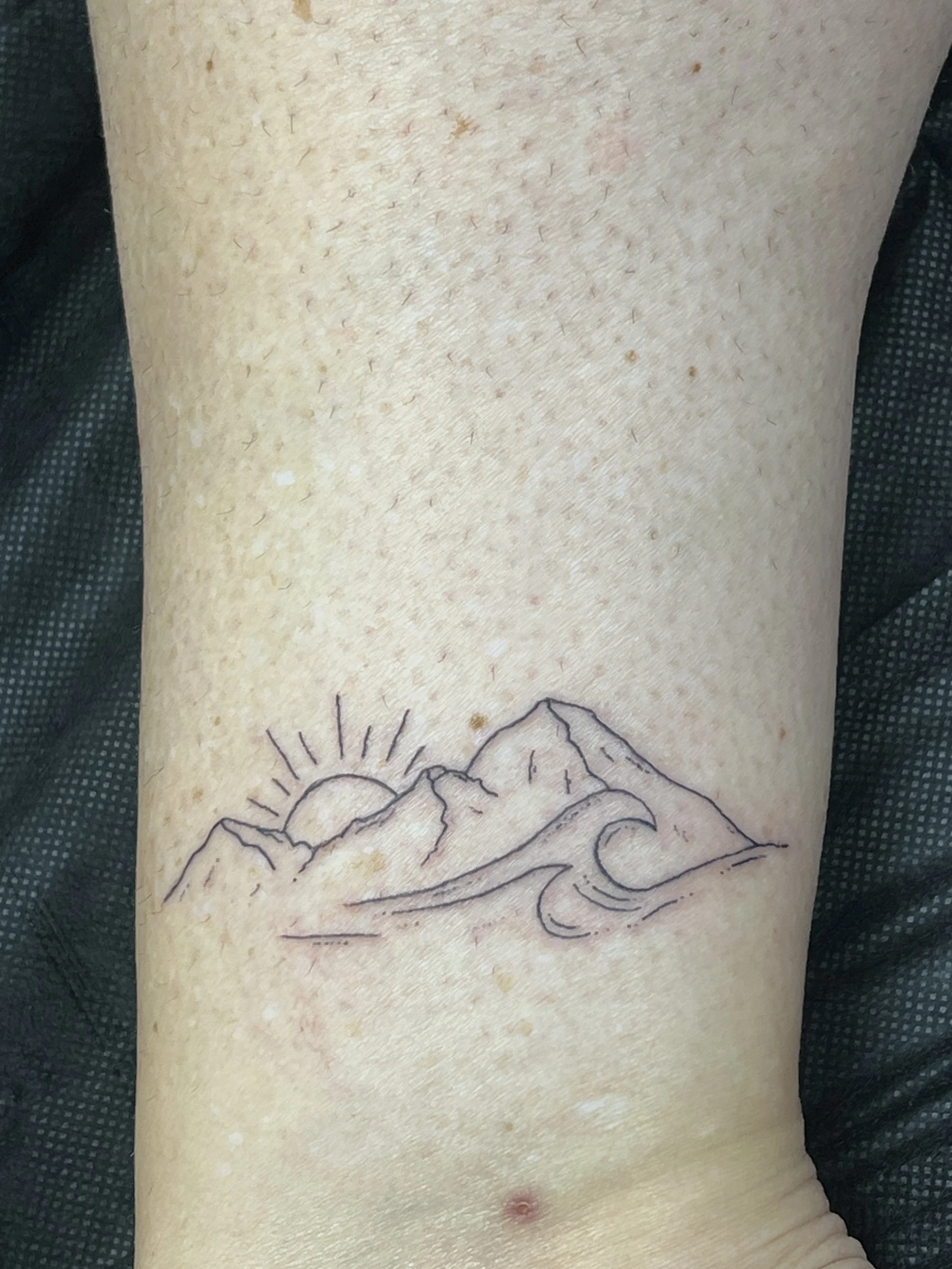A close-up of a person's side torso with a black outline tattoo of mountains, a sun, and waves on their skin.