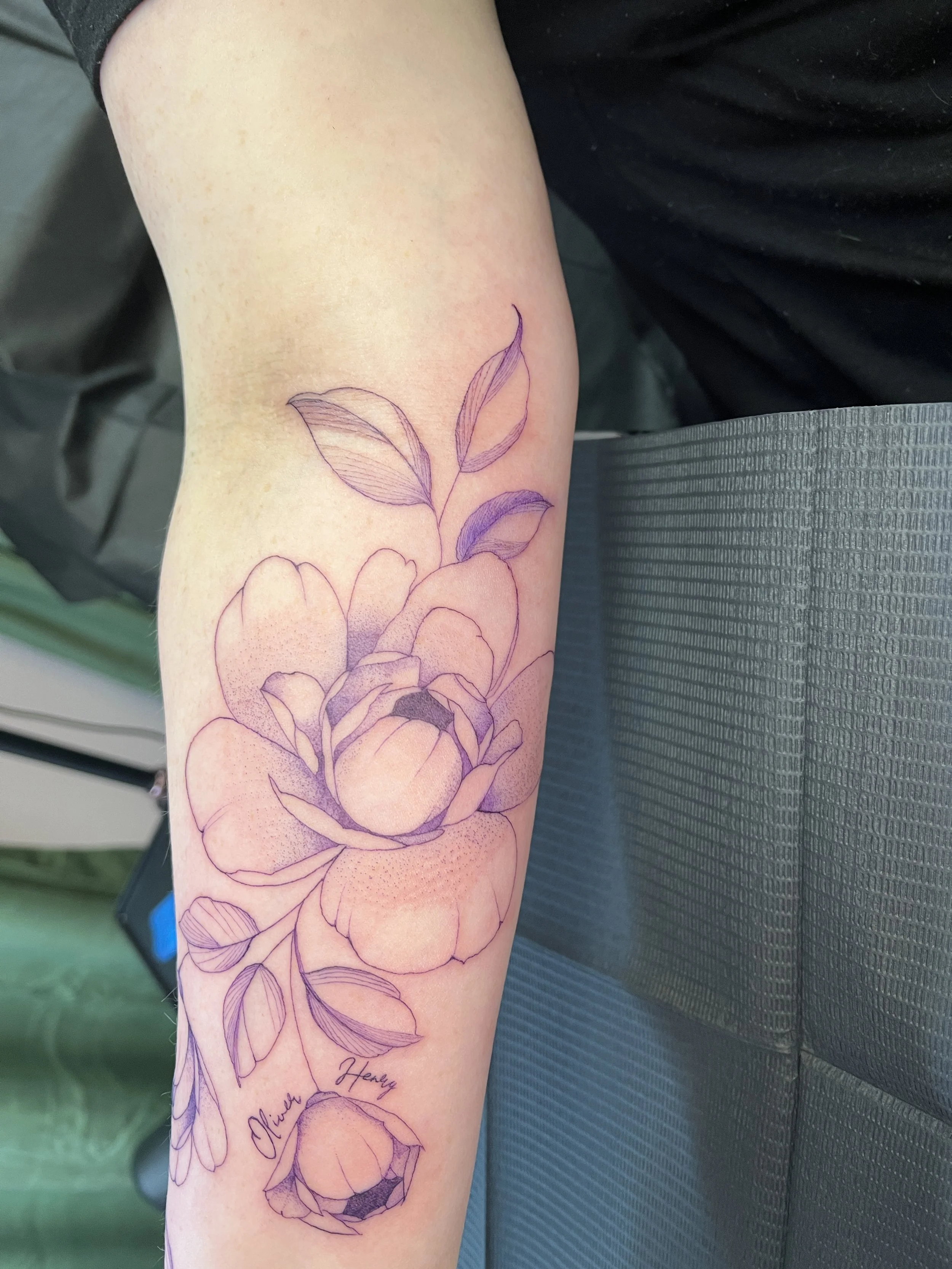 A tattoo of a flower with multiple petals and leaves, sketched in purple, on a person's forearm.
