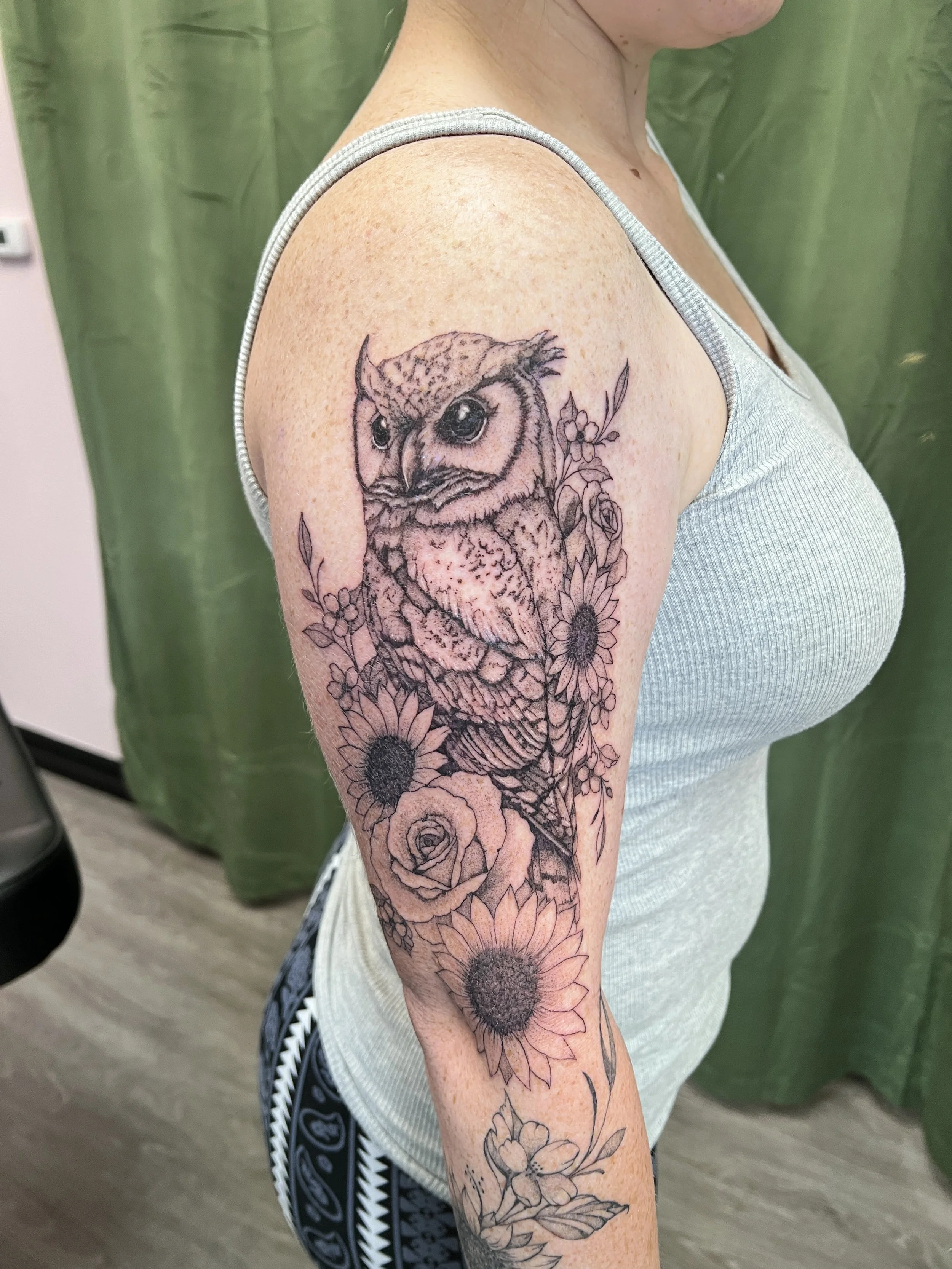 A person with a black and gray tattoo of an owl surrounded by flowers on their upper arm.