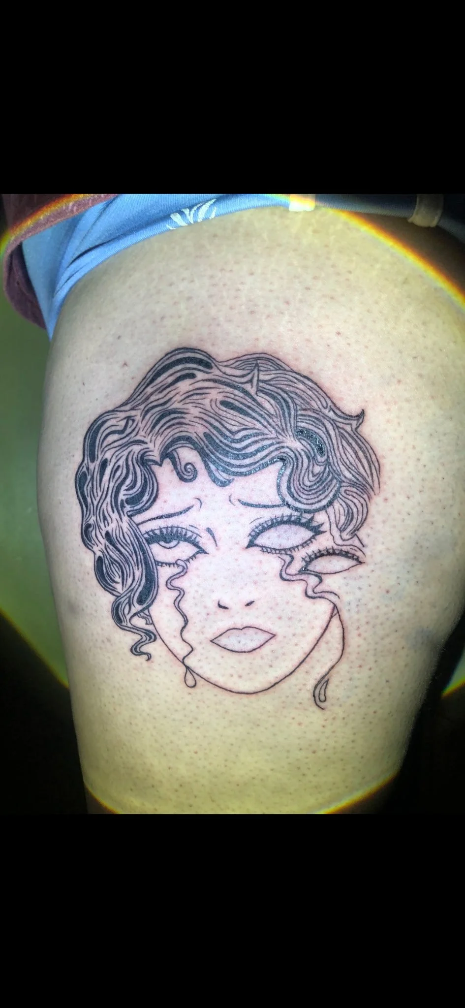Tattoo of a woman's face with wavy hair and tears, on the thigh of a person wearing blue and yellow clothing.